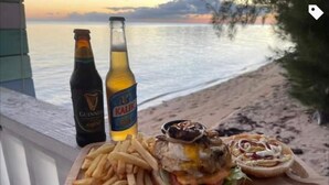 A wooden table with a burger, fries, and two beer bottles on a balcony overlooking a beach. - Coral Harbour Beach House And Villas (Nassau)
