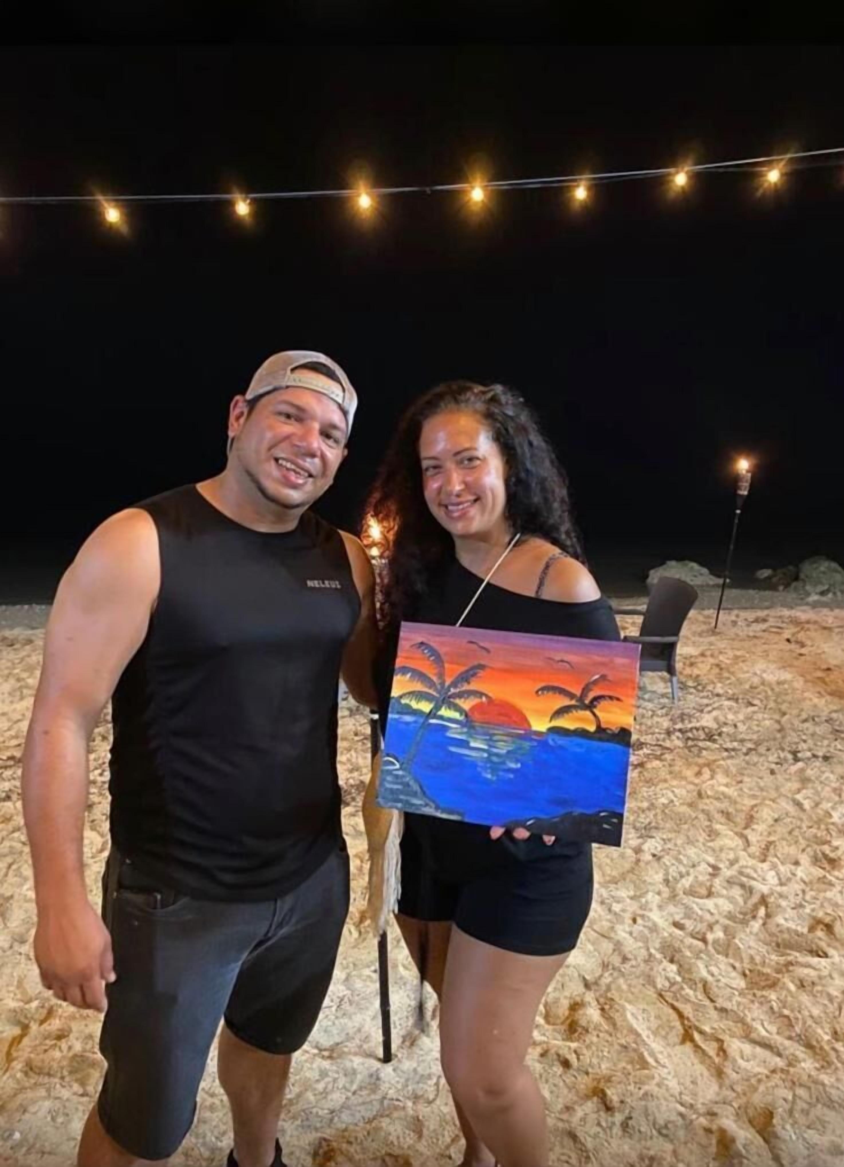 Dos personas de pie en una playa arenosa de noche, con luces colgantes arriba.