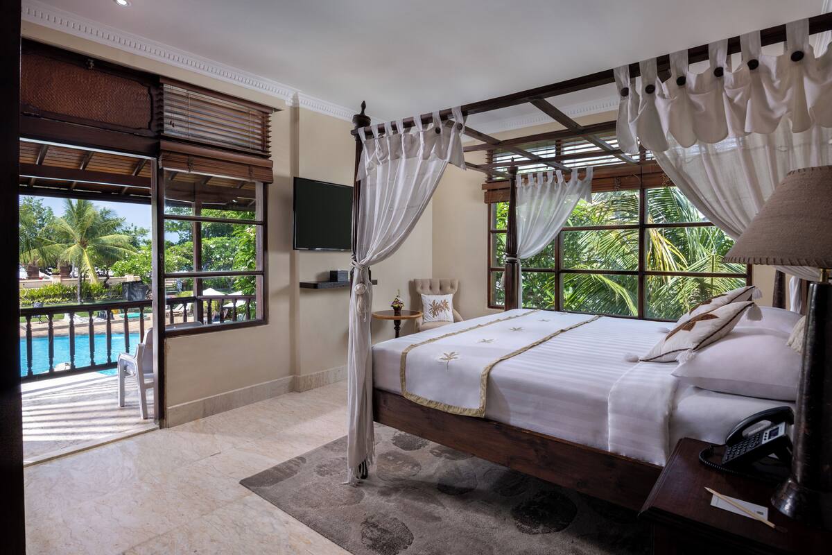 A bedroom with a large bed, a bedside table with a lamp, a TV mounted on the wall, and a view of a pool and palm trees.