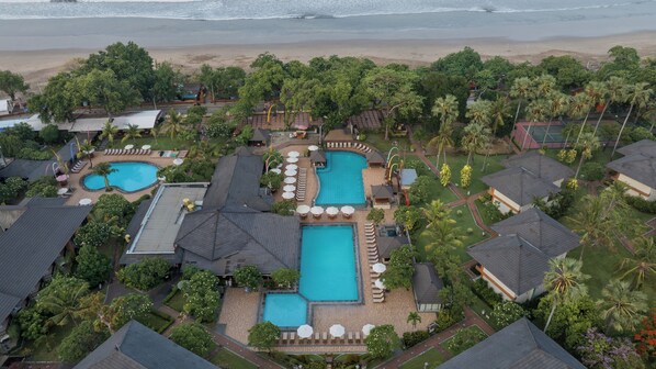 An aerial view of a resort with multiple swimming pools, lush greenery, and a beach in the background. - The Jayakarta Bali (Legian)