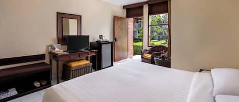 A hotel room with a large bed, a desk with a television, a small refrigerator, and a window with a view of greenery.