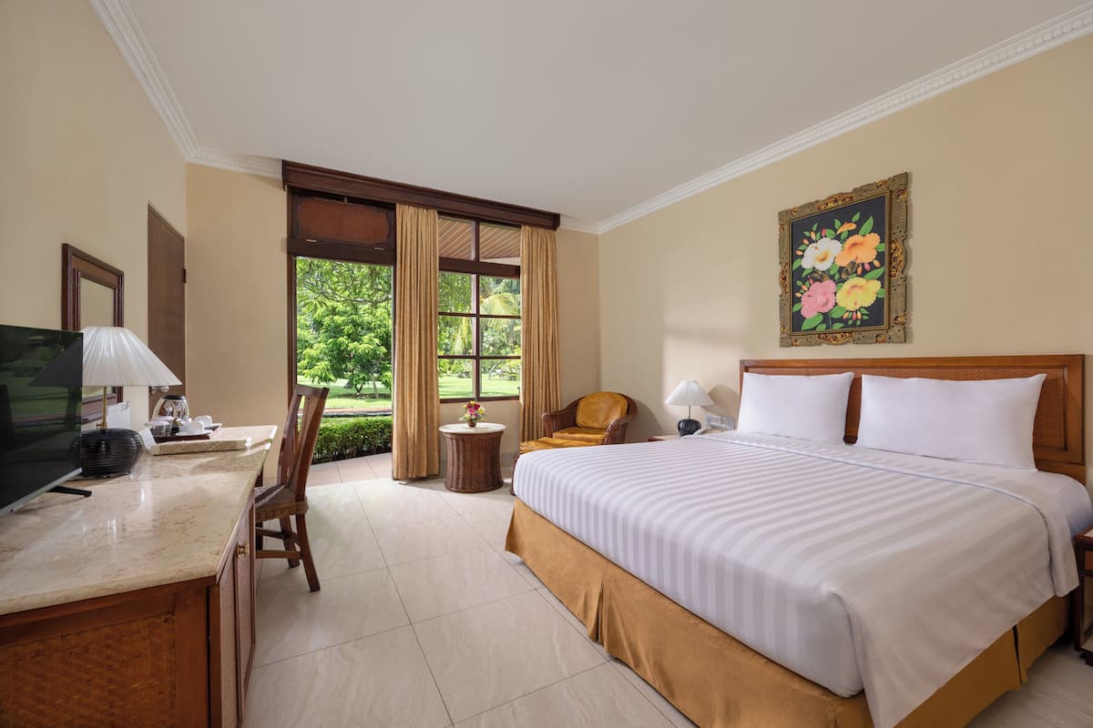 A hotel room with a bed, a desk with a TV, a chair, a small table with a lamp, and a painting on the wall.