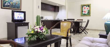 Una sala de estar con un sofá, una mesa de centro con un arreglo floral, un televisor sobre un mueble, una zona de comedor con sillas y una mesa, y una cocina al fondo.