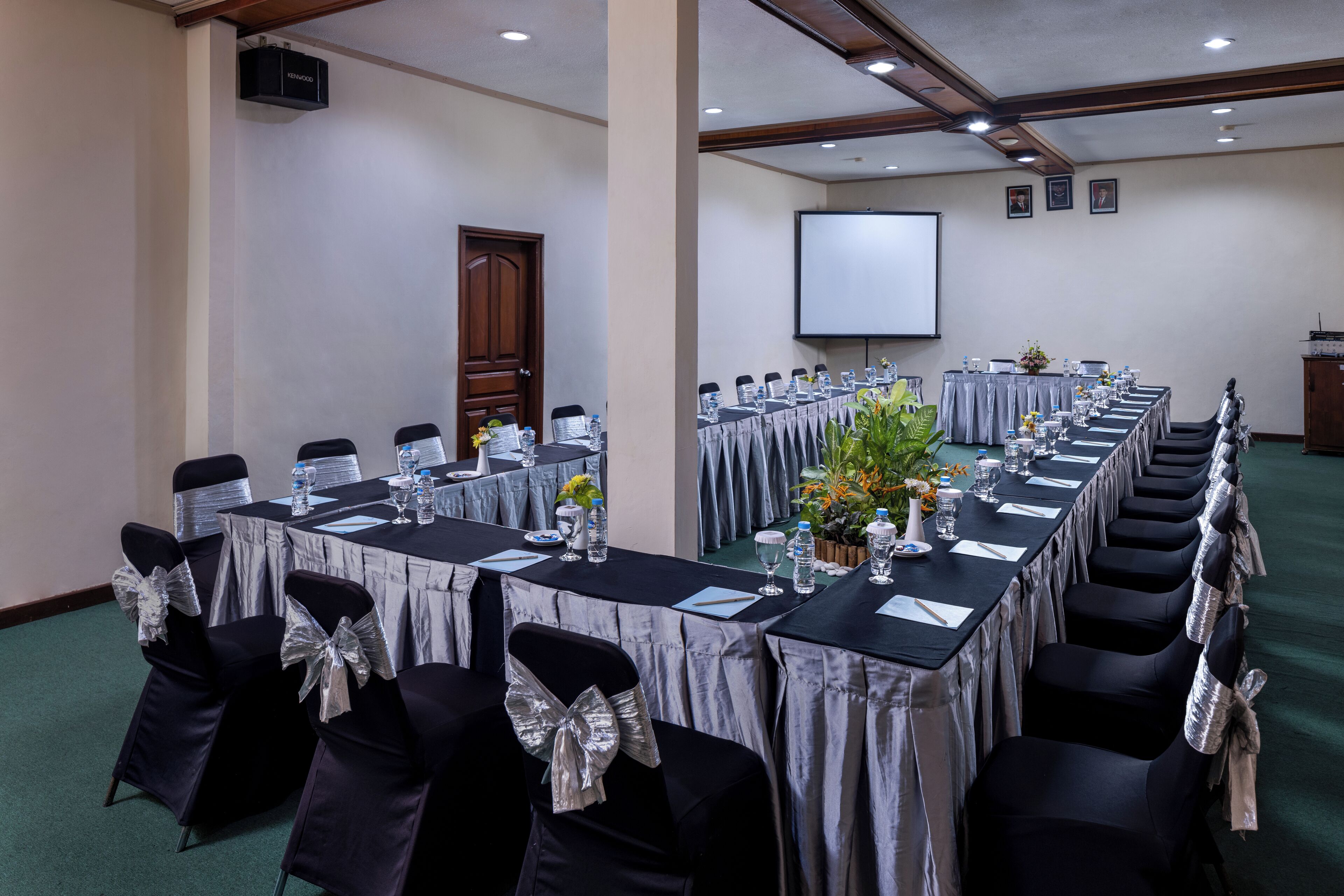 A conference room with long tables, chairs, a projector screen, and a screen on the wall.