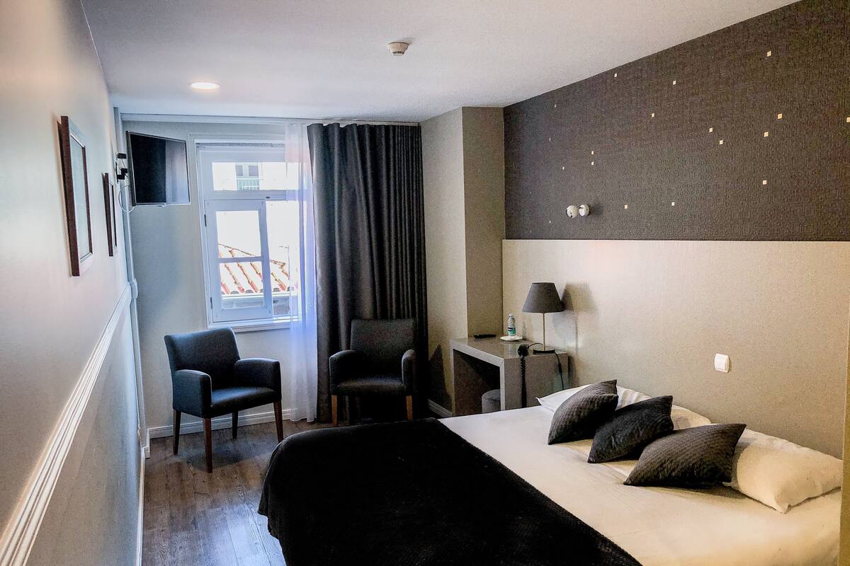 A hotel room with a bed, two chairs, a desk with a lamp, and a window with curtains.
