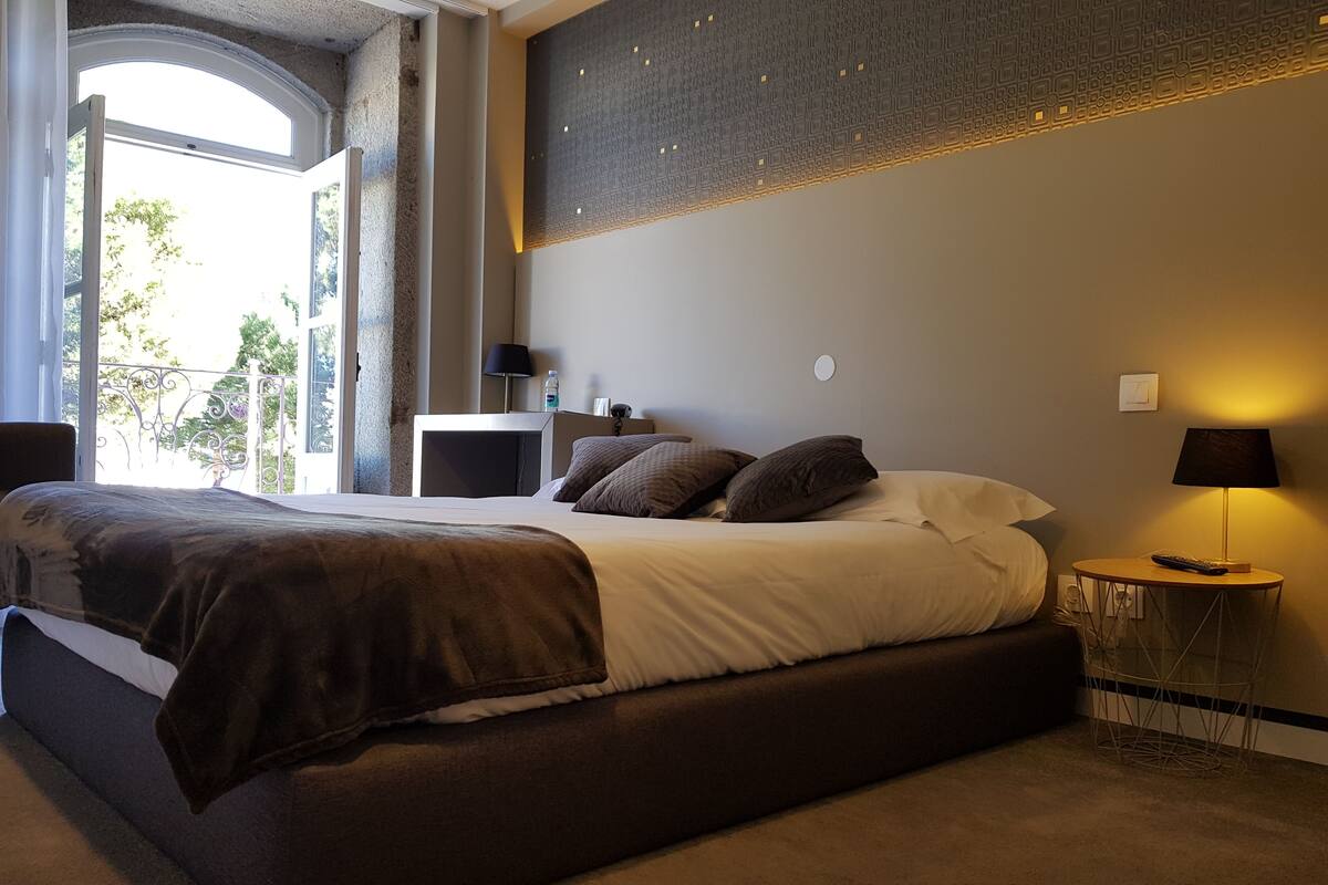 A modern bedroom with a large bed, bedside tables, a lamp, and a view of greenery outside.