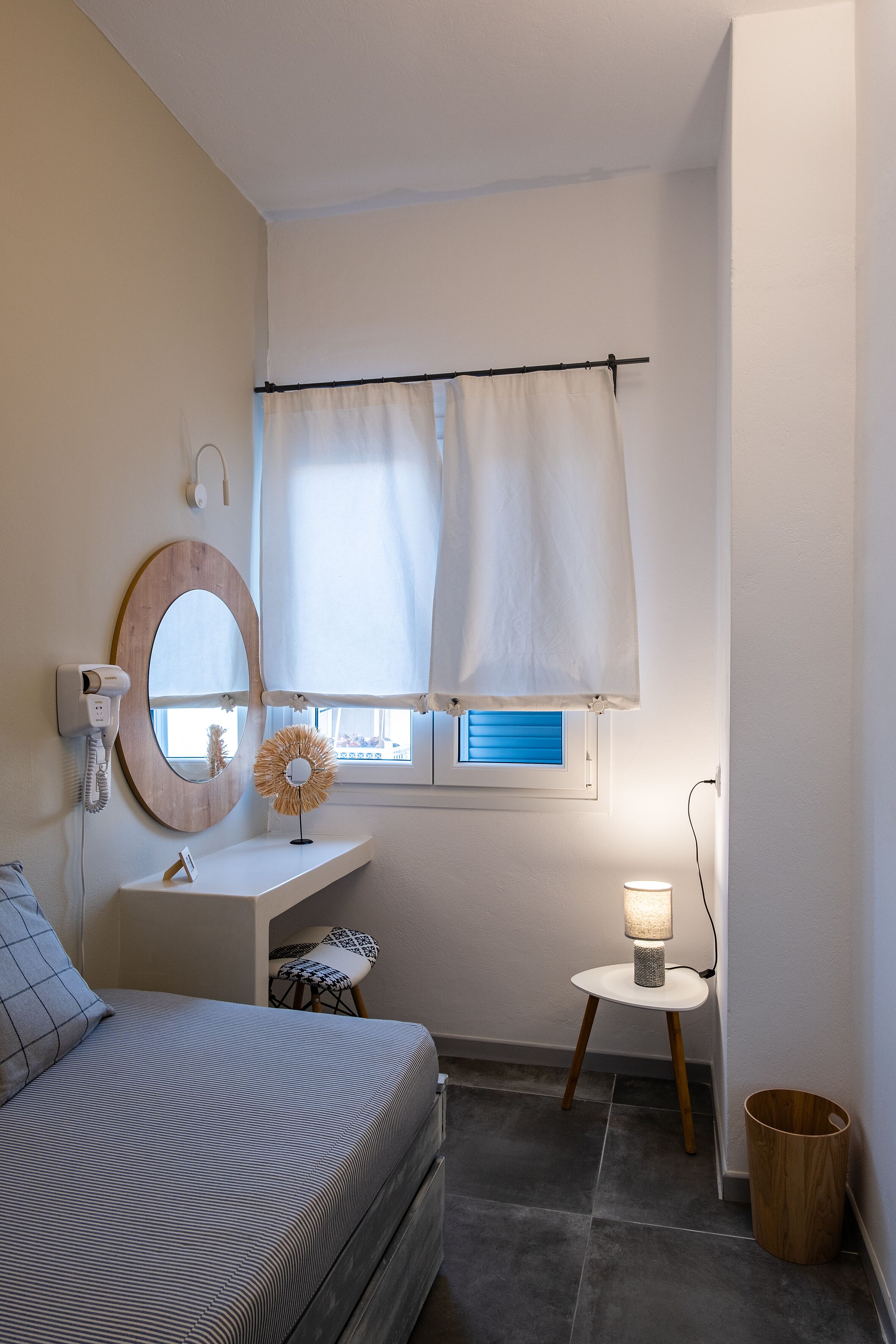 Un dormitorio con cama, mesita de noche, espejo, ventana con cortinas, lámpara y un taburete pequeño.
