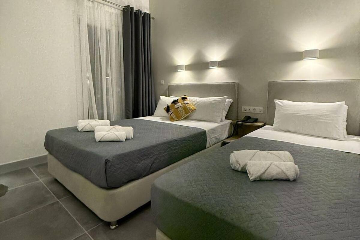 A hotel room with two beds, grey bedding, a small table with a phone, and a window with curtains.