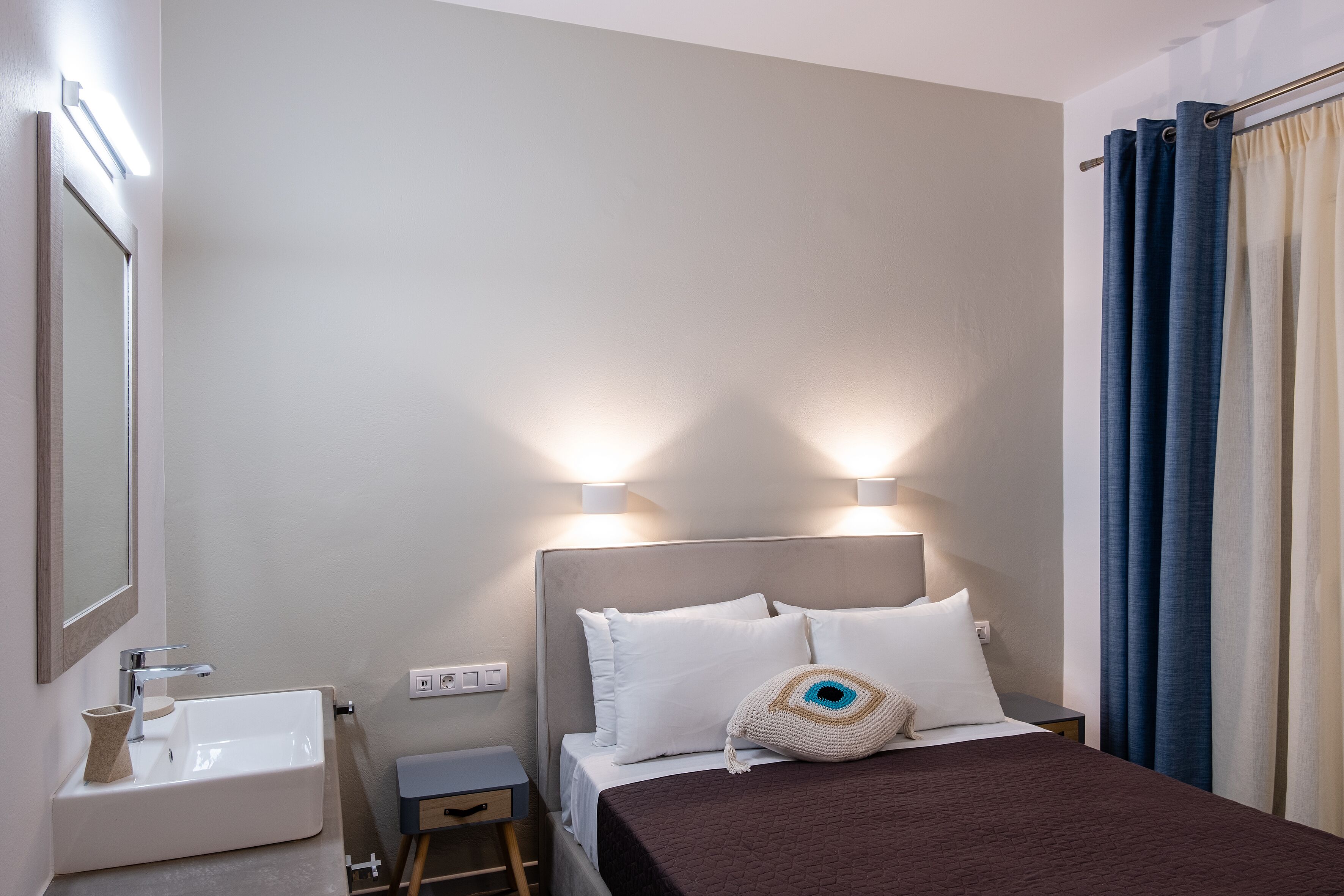 Habitación de hotel con cama, mesita de noche, lámpara, espejo, lavamanos y cortinas.