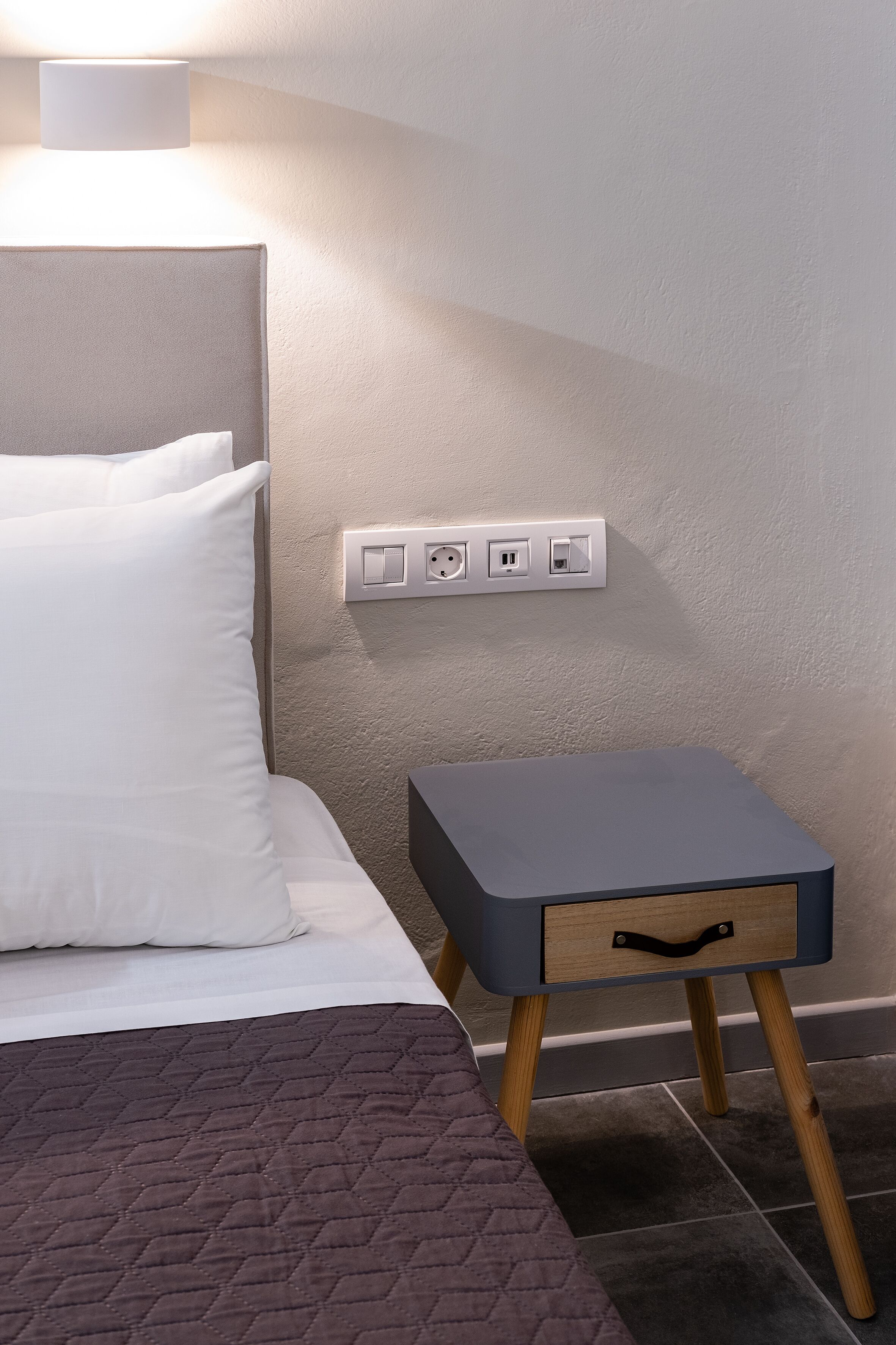 A neatly made bed with a white pillow and a dark quilted blanket. A small bedside table with a drawer is positioned next to the bed.