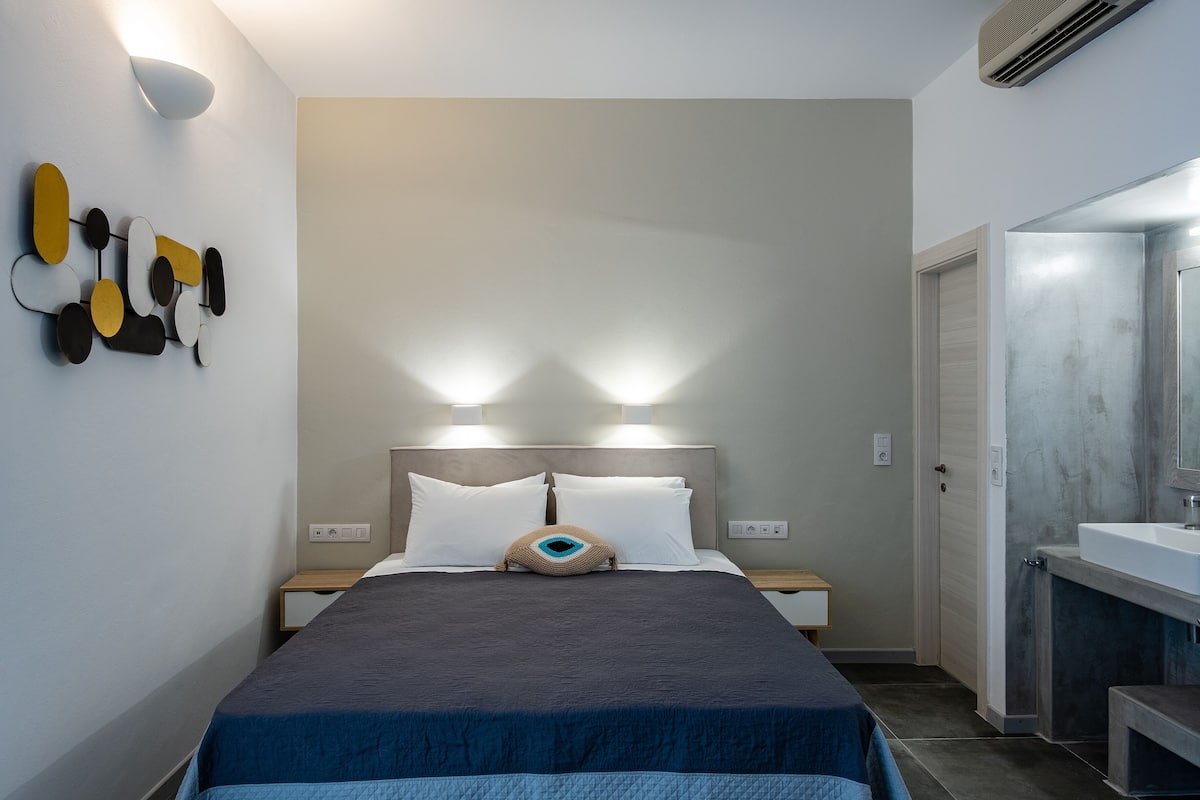 A modern hotel room with a neatly made bed, a bedside table, a wall-mounted light fixture, and a bathroom with a sink and mirror.