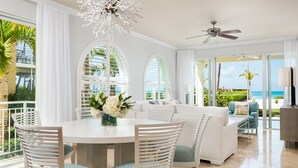 A modern living room with a white sofa, a dining table, and chairs. There are large windows with white curtains, a ceiling fan, and a view of the ocean.