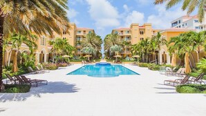 A resort pool area with multiple lounge chairs and umbrellas, surrounded by palm trees and a building with balconies.