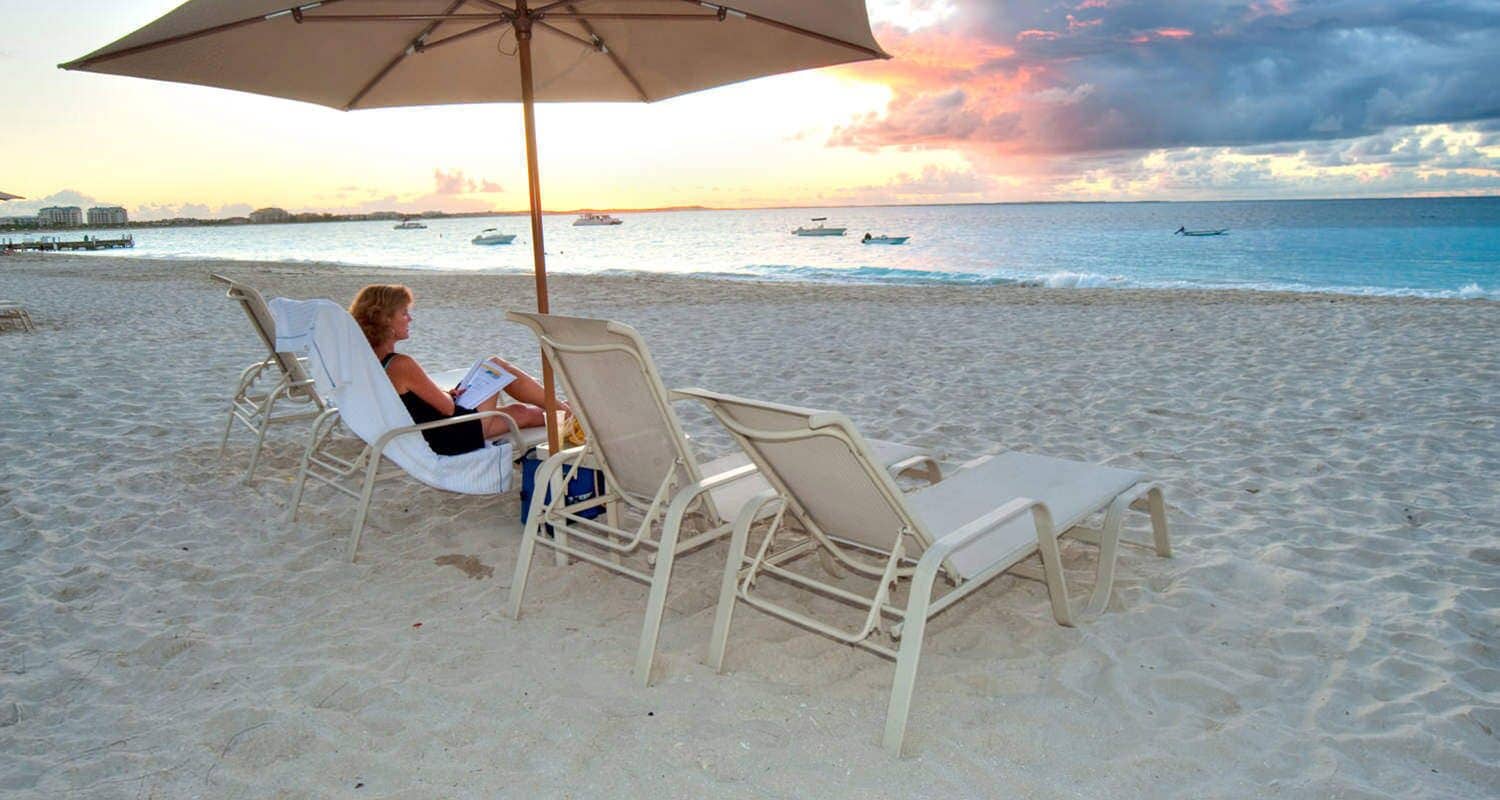 A beach with two lounge chairs under a large umbrella, a person reading a book, and boats in the water.