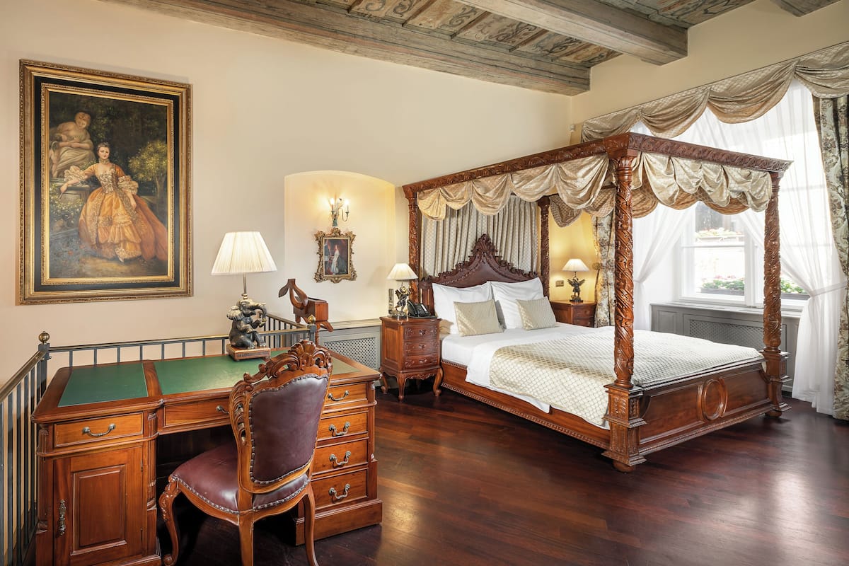 Una cama con dosel y cuatro columnas, un escritorio de fieltro verde y una silla de cuero, un cuadro grande enmarco y dos mesitas de noche con lámparas.