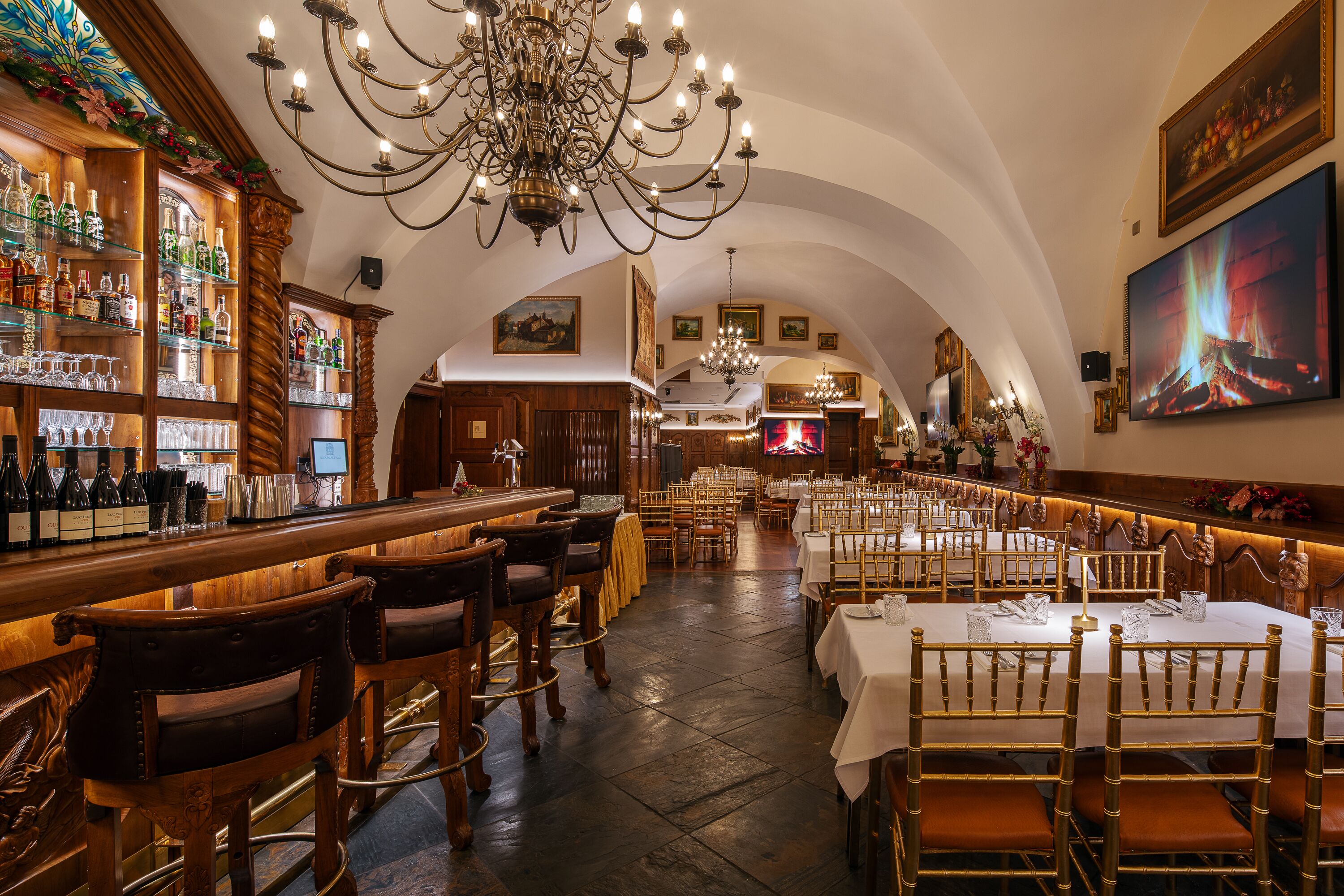 A restaurant with a bar area, wooden bar stools, tables with white tablecloths, and a chandelier.