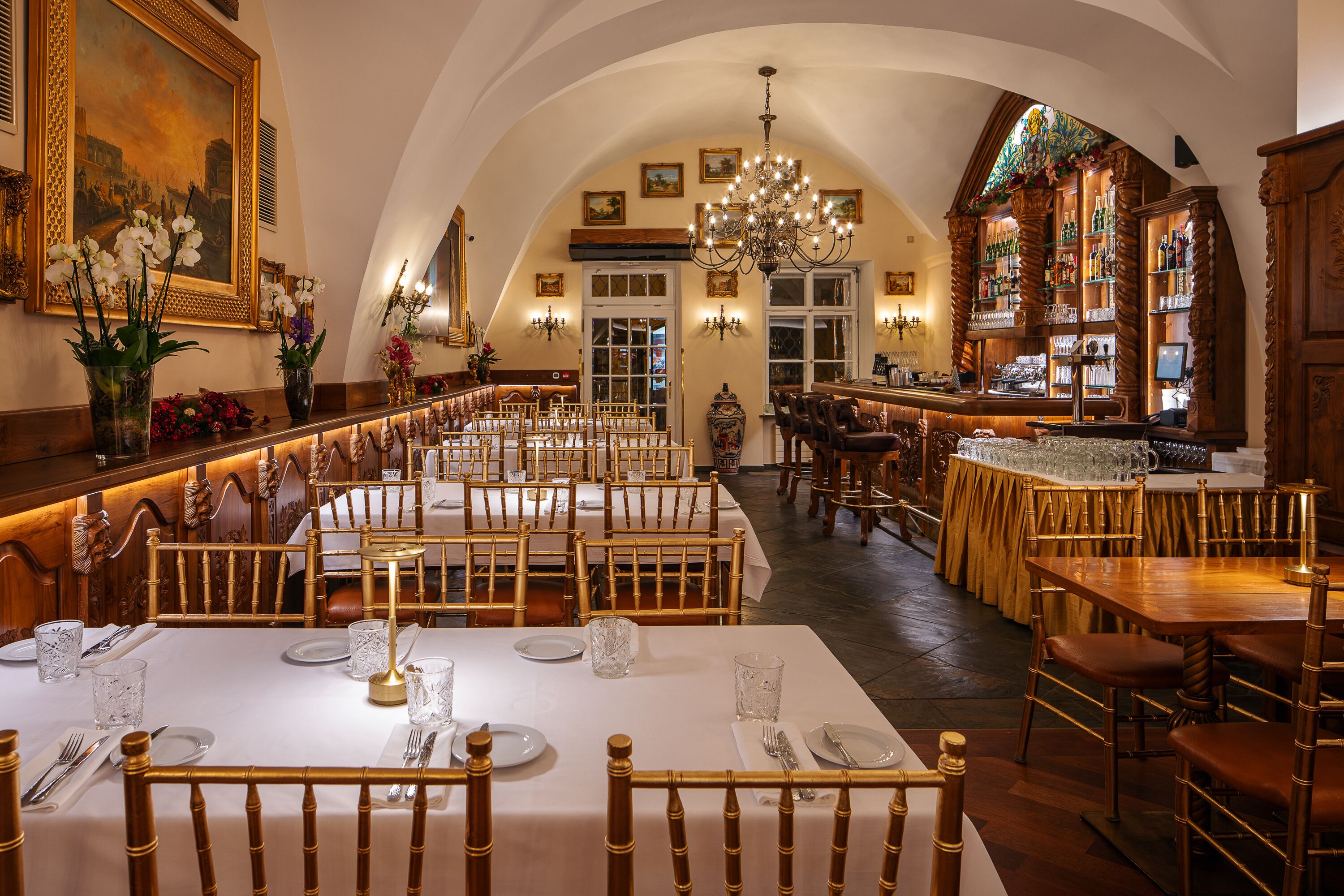 A restaurant with multiple dining tables and chairs, a bar area, and a chandelier.