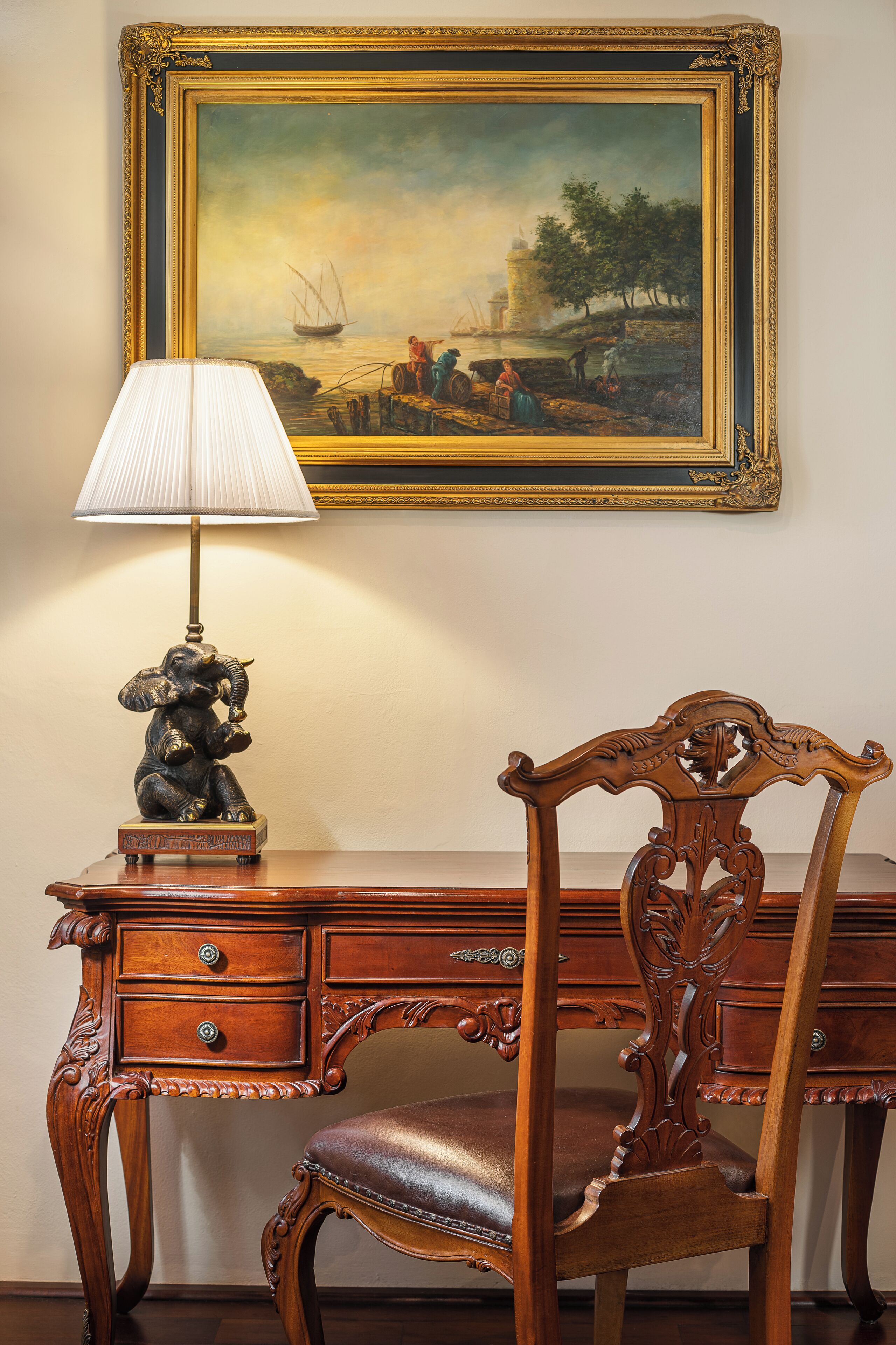 A wooden desk with a lamp and a chair, a framed painting above the desk, and a small elephant statue on the desk.