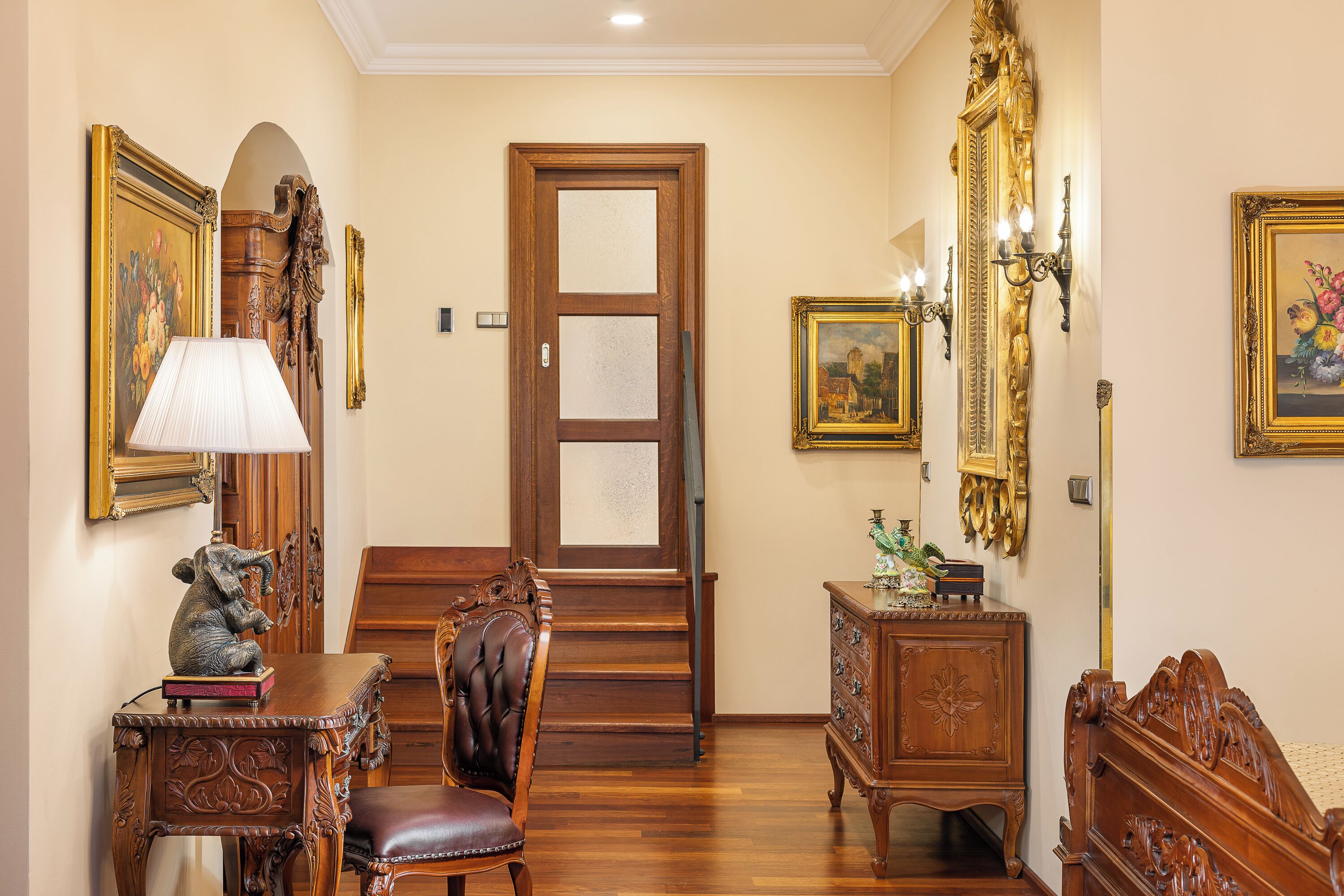 Une pièce avec un plancher en bois, une chaise en bois, un bureau en bois avec une lampe, une commode et une porte dotée d’un panneau en verre dépoli.