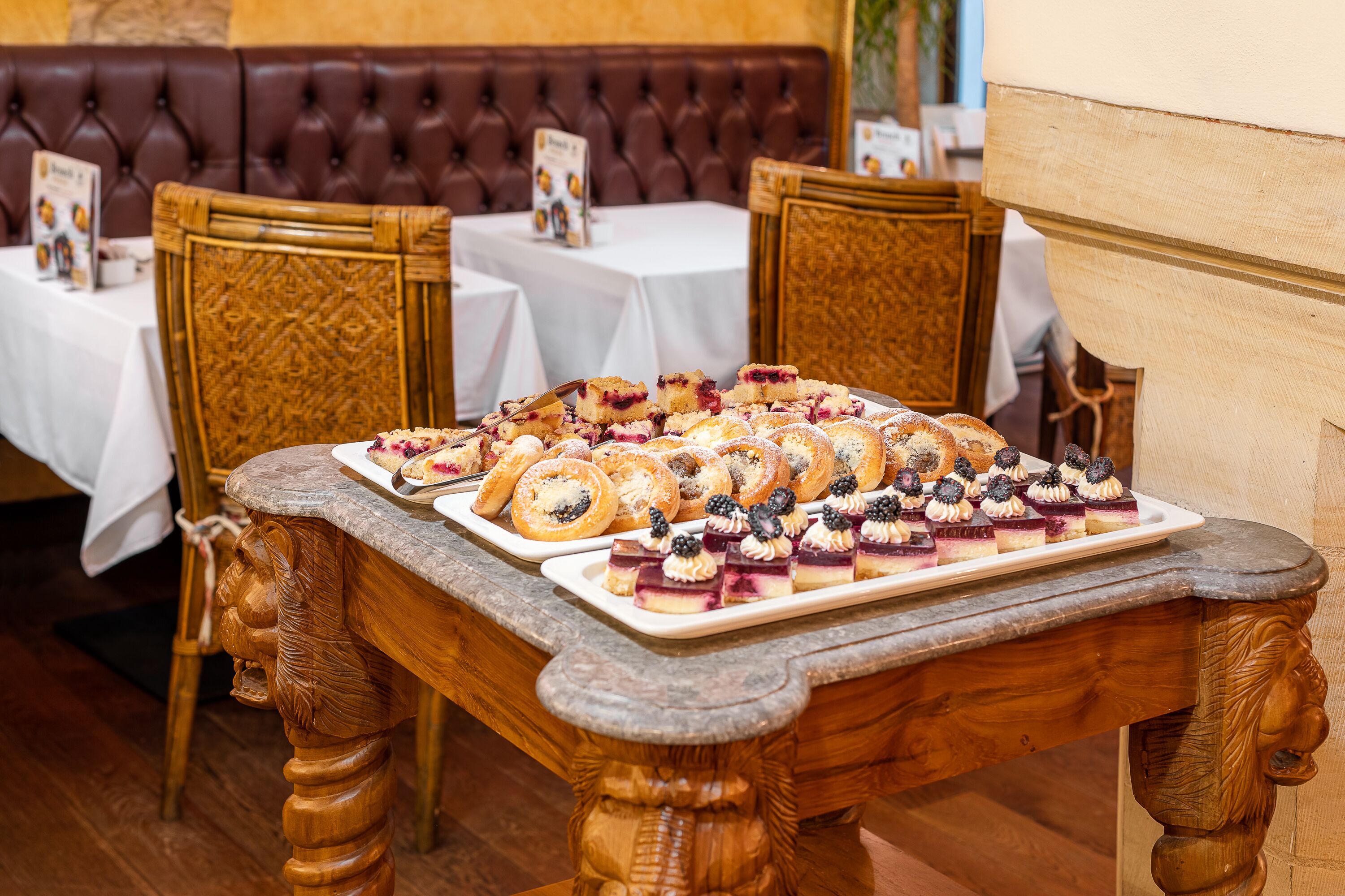 Una mesa de buffet con diversos postres, entre ellos pasteles y productos de pastelería, en un restaurante con sillas de madera y un asiento tapizado de cuero al fondo.