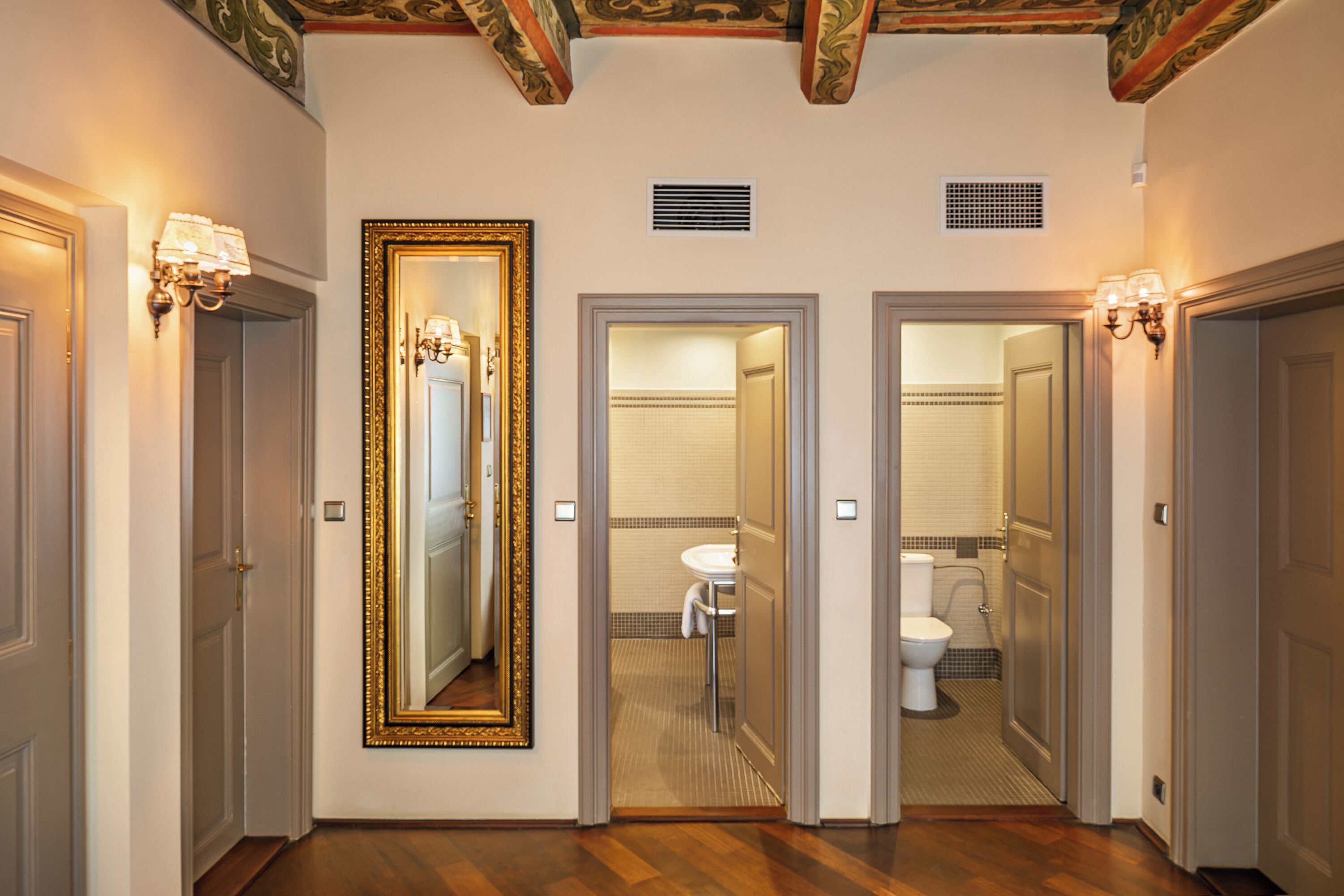 Un pasillo con un espejo grande, dos puertas que dan a un baño y una lámpara en la pared.