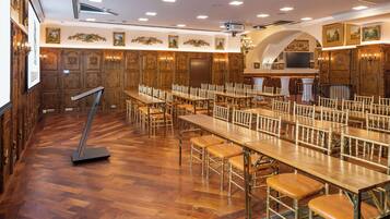 Un'ampia sala da pranzo con lunghe tavole e sedie in legno, uno schermo per proiezioni e una scala che conduce a un piano superiore.