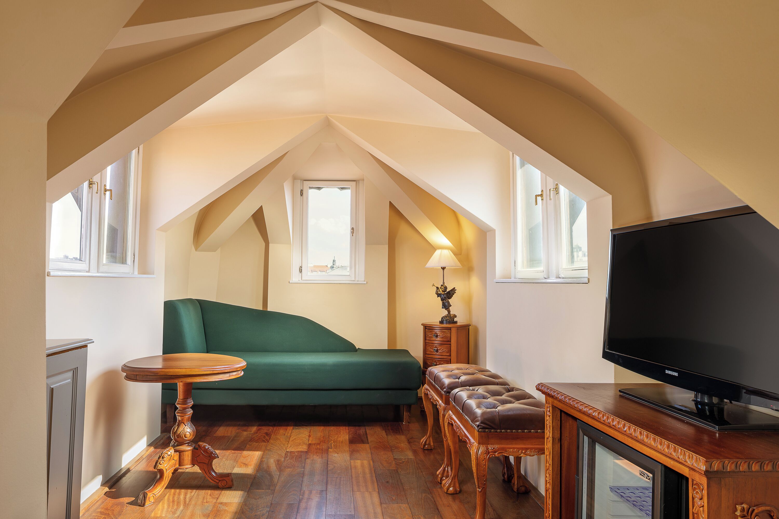 A cozy attic room with a green sofa, a wooden table, a leather ottoman, and a TV on a wooden stand.