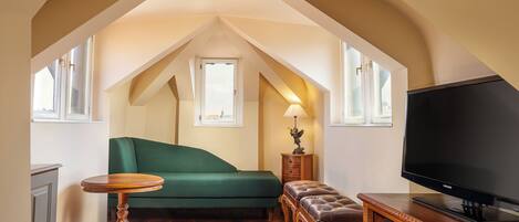 A cozy attic room with a green sofa, a wooden table, a leather ottoman, and a TV on a wooden stand.
