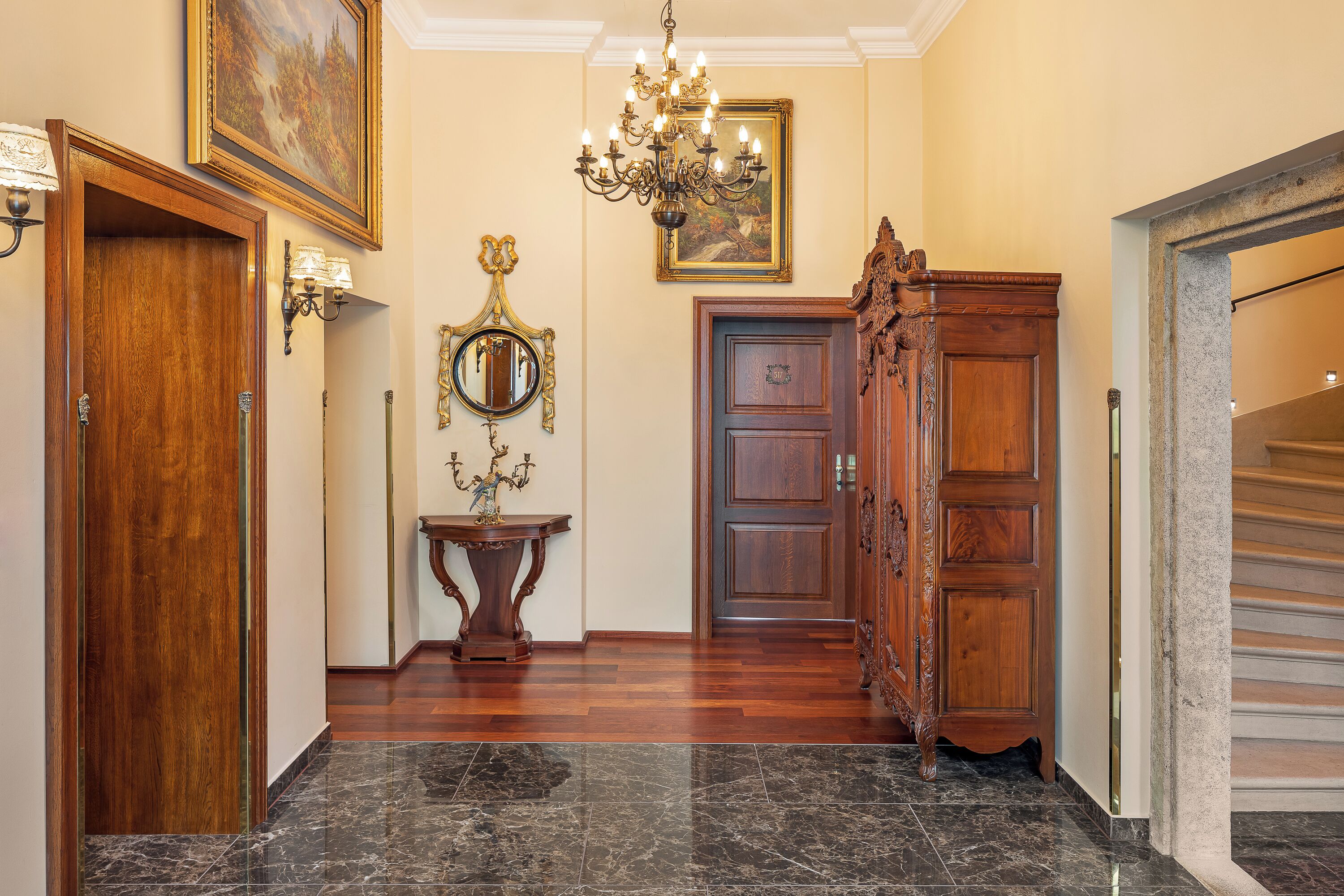 Um corredor com piso de mármore, portas de madeira, um candelabro e obras de arte nas paredes.