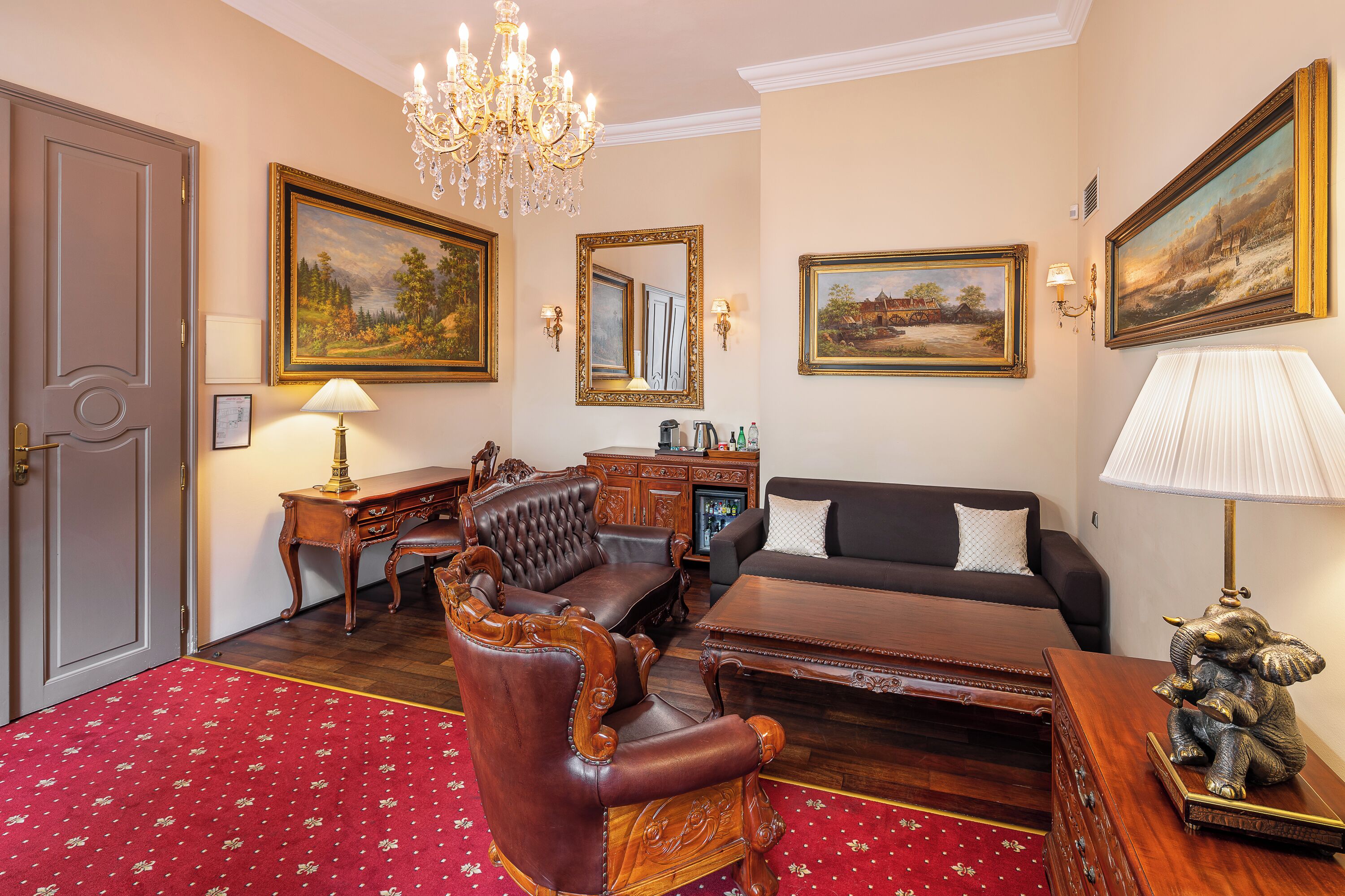 A room with a wooden desk, leather armchair, and a sofa. There are paintings on the walls, a chandelier, and a red carpet.