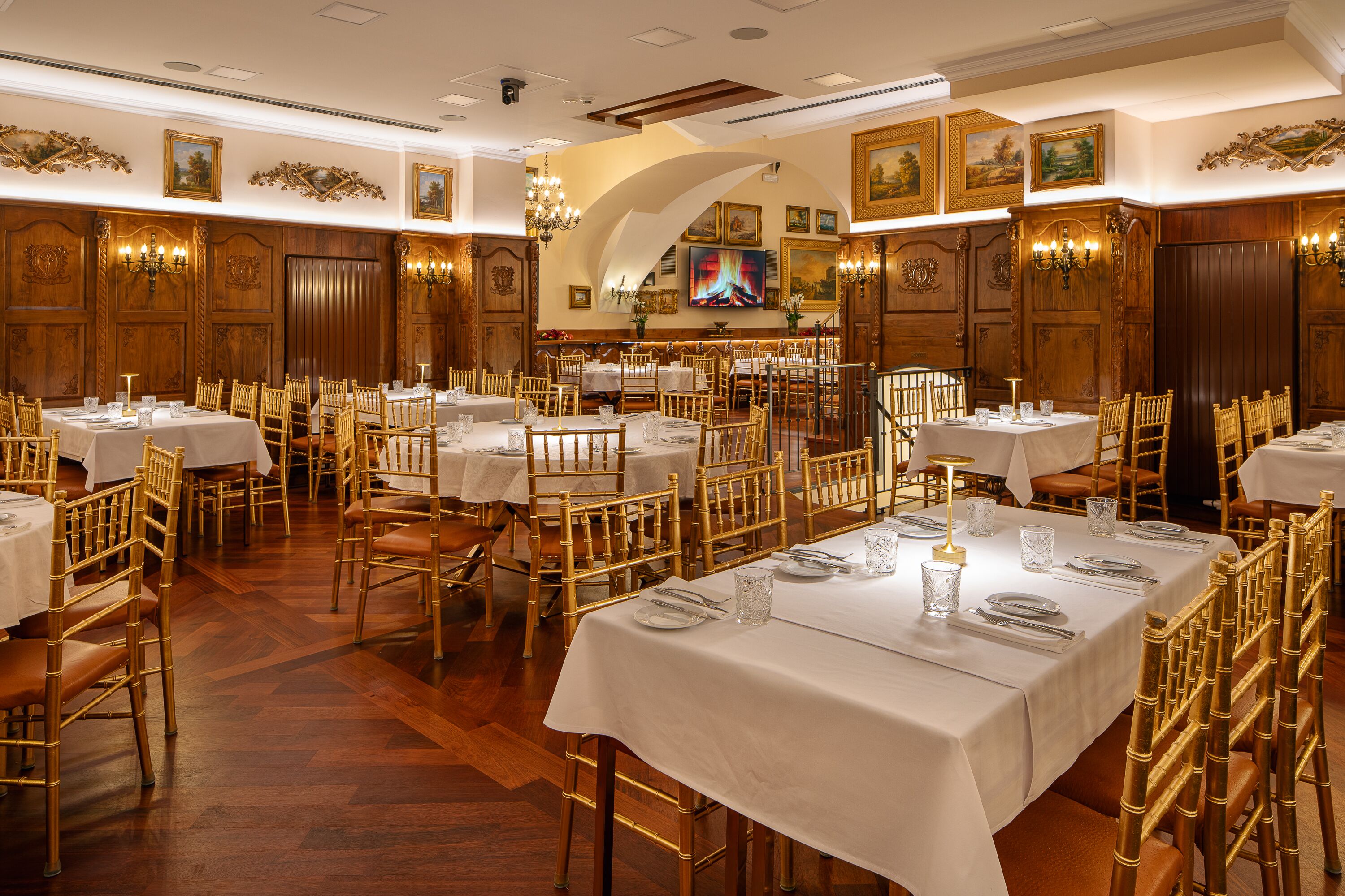 Comedor formal con varias mesas dispuestas para el servicio, paneles de madera y una pantalla de televisión en la pared.