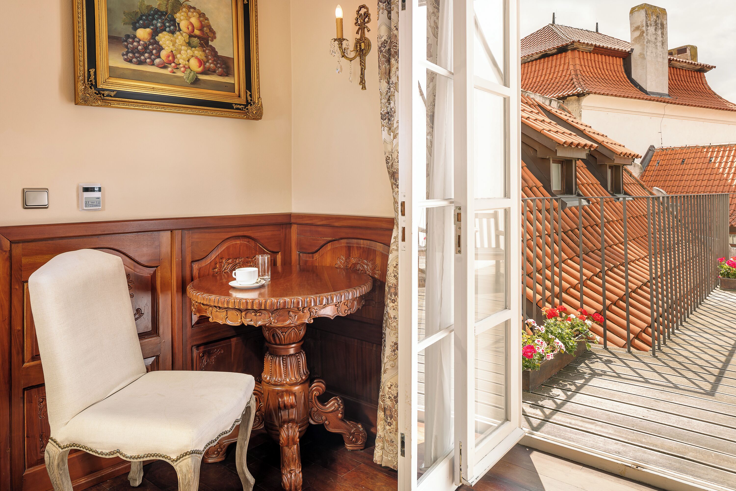A corner room with a wooden chair, a table, a painting, and a balcony with a tiled roof and flowers.