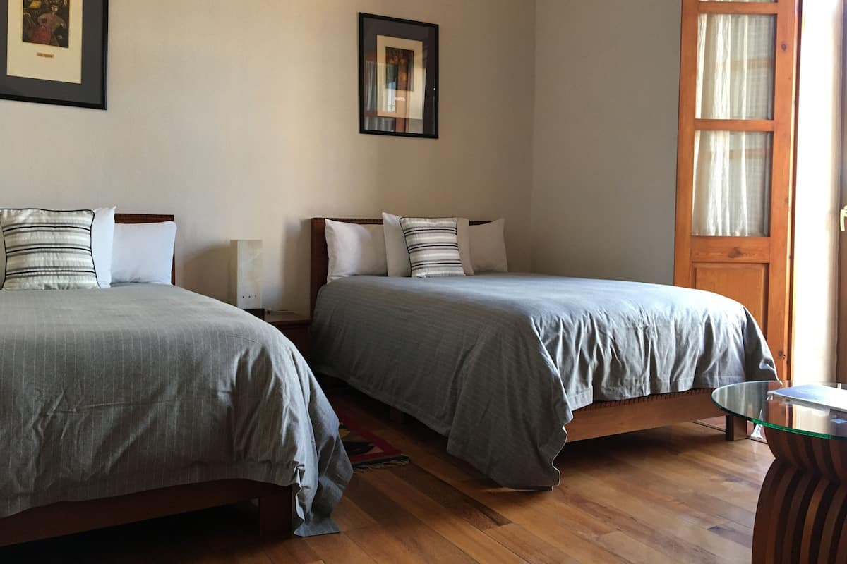 Quarto com duas camas, piso de madeira, mesa de vidro e porta com cortinas.