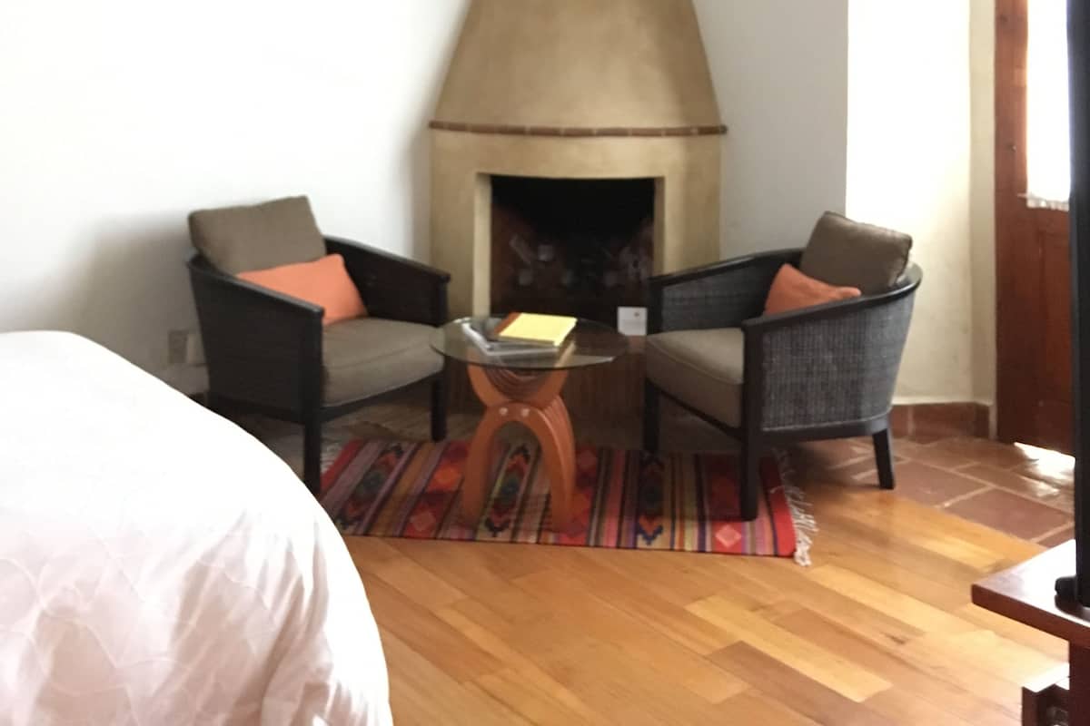 Um quarto com lareira, duas poltronas, uma mesa pequena e uma cama.