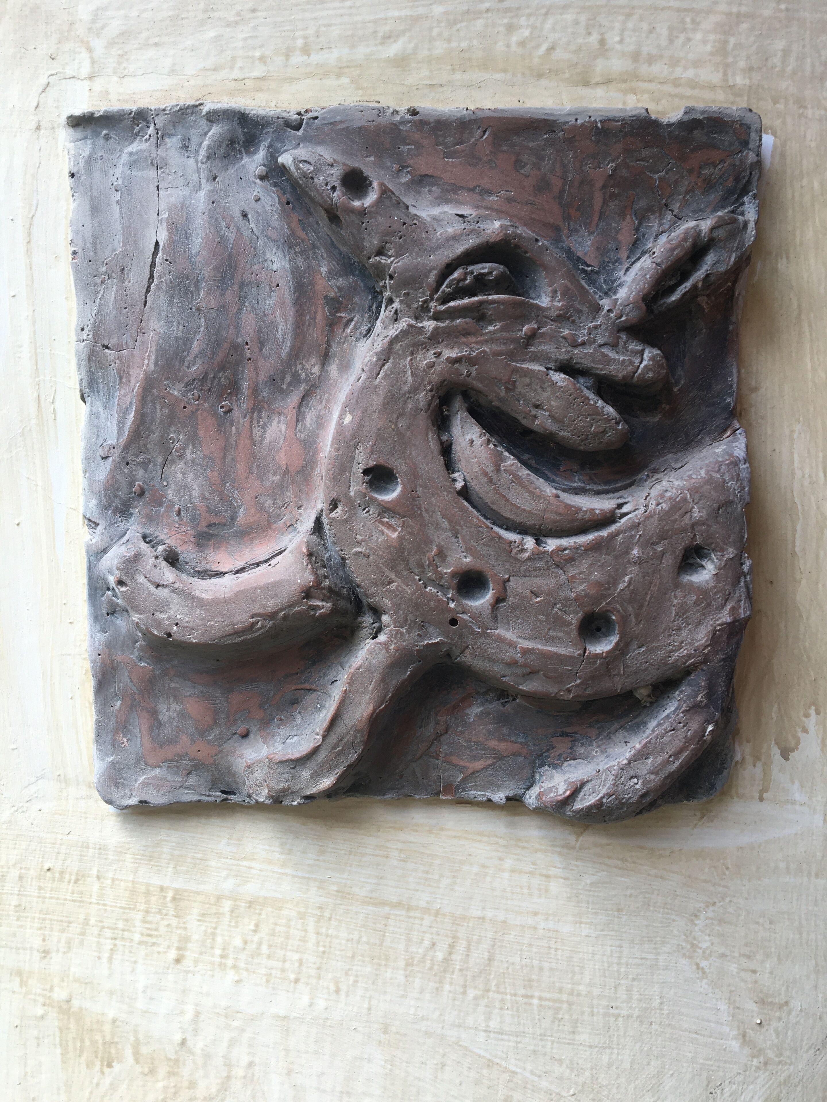 Una escultura de piedra oscura y texturizada con un diseño en espiral y agujeros, montada sobre una pared lisa.
