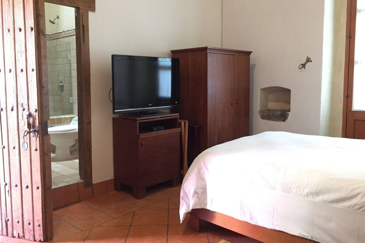 Quarto com cama, um armário de madeira, televisão e banheiro visível por uma porta aberta.