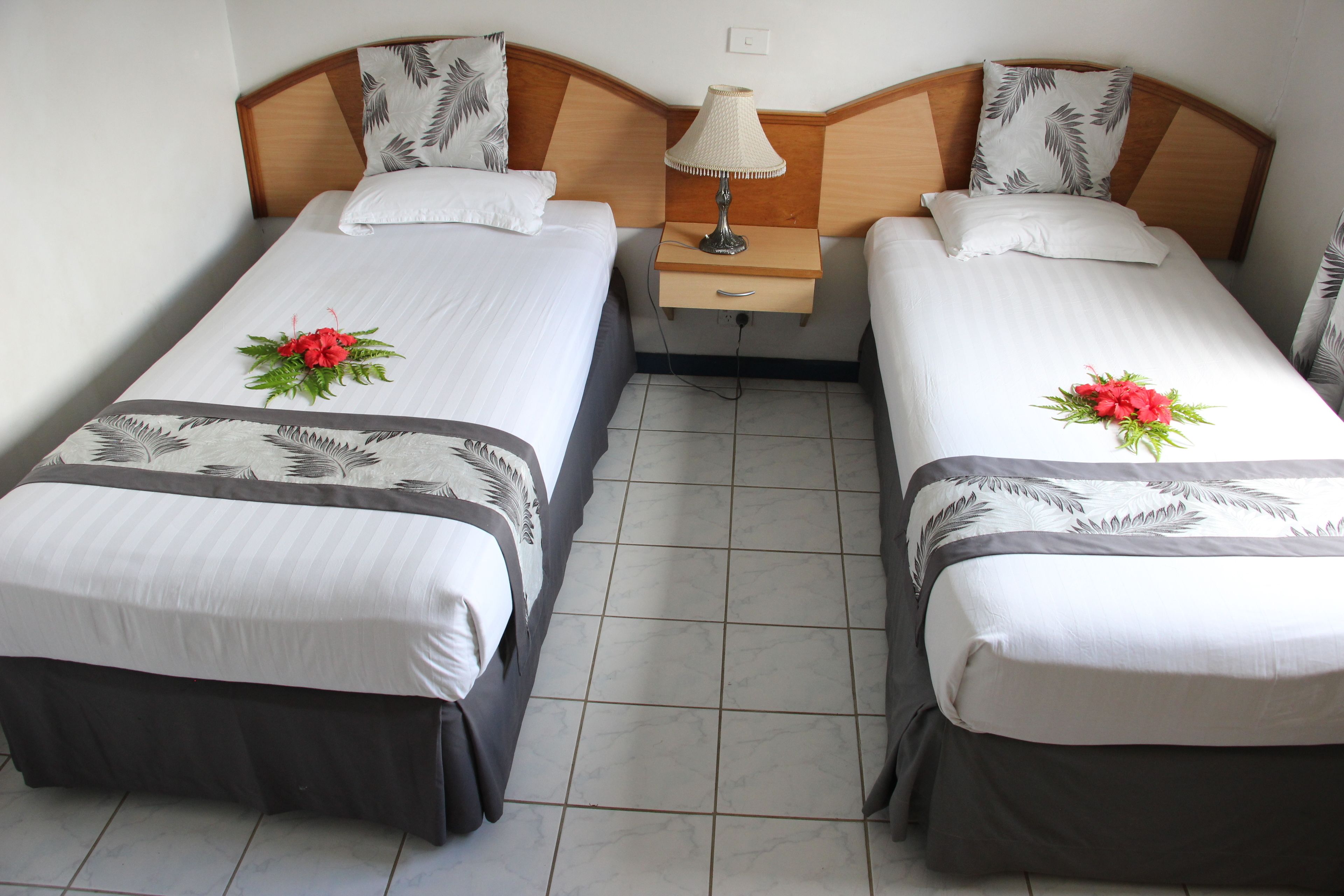 Habitación de hotel con dos camas individuales, cada una con un arreglo floral, una mesita de noche con lámpara y cabecero de madera.