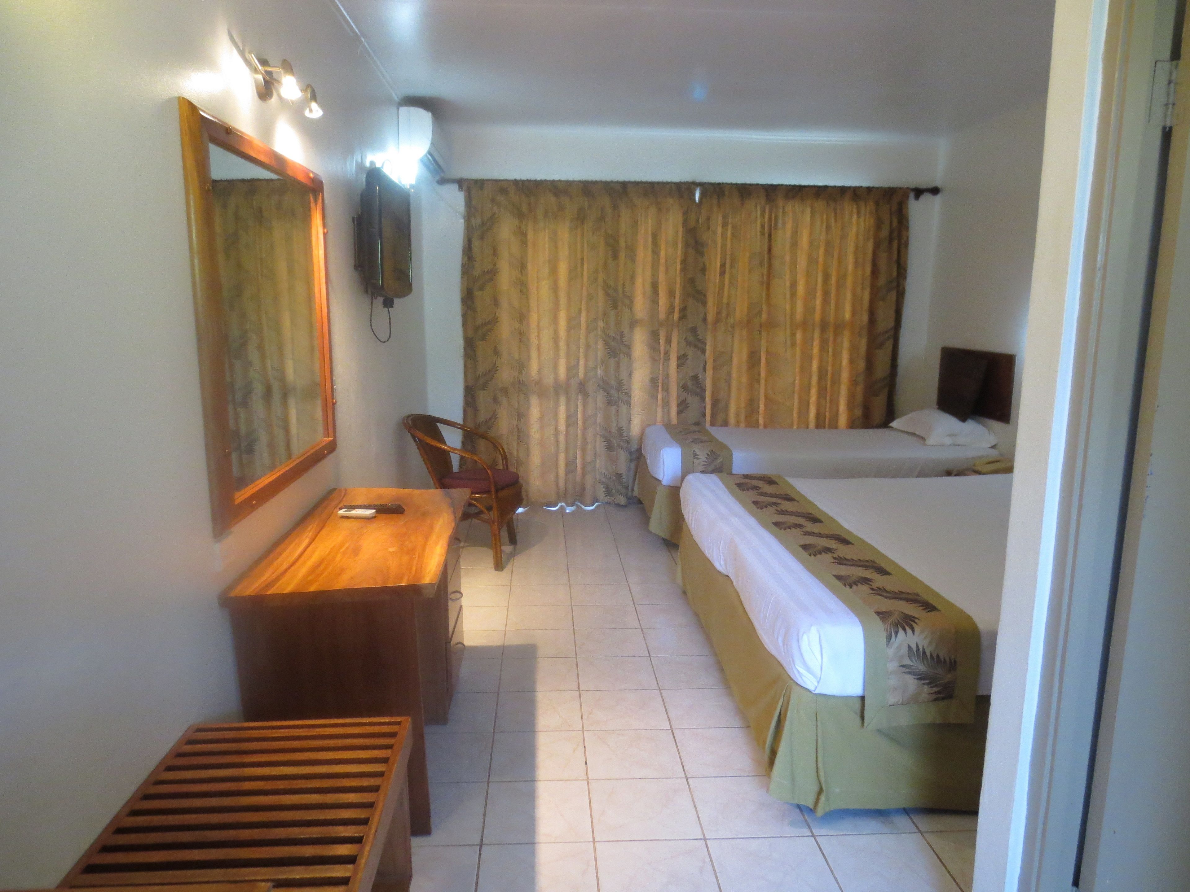 Habitación de hotel con un escritorio de madera, una silla, una cama con colcha verde, un espejo grande y una cortina.