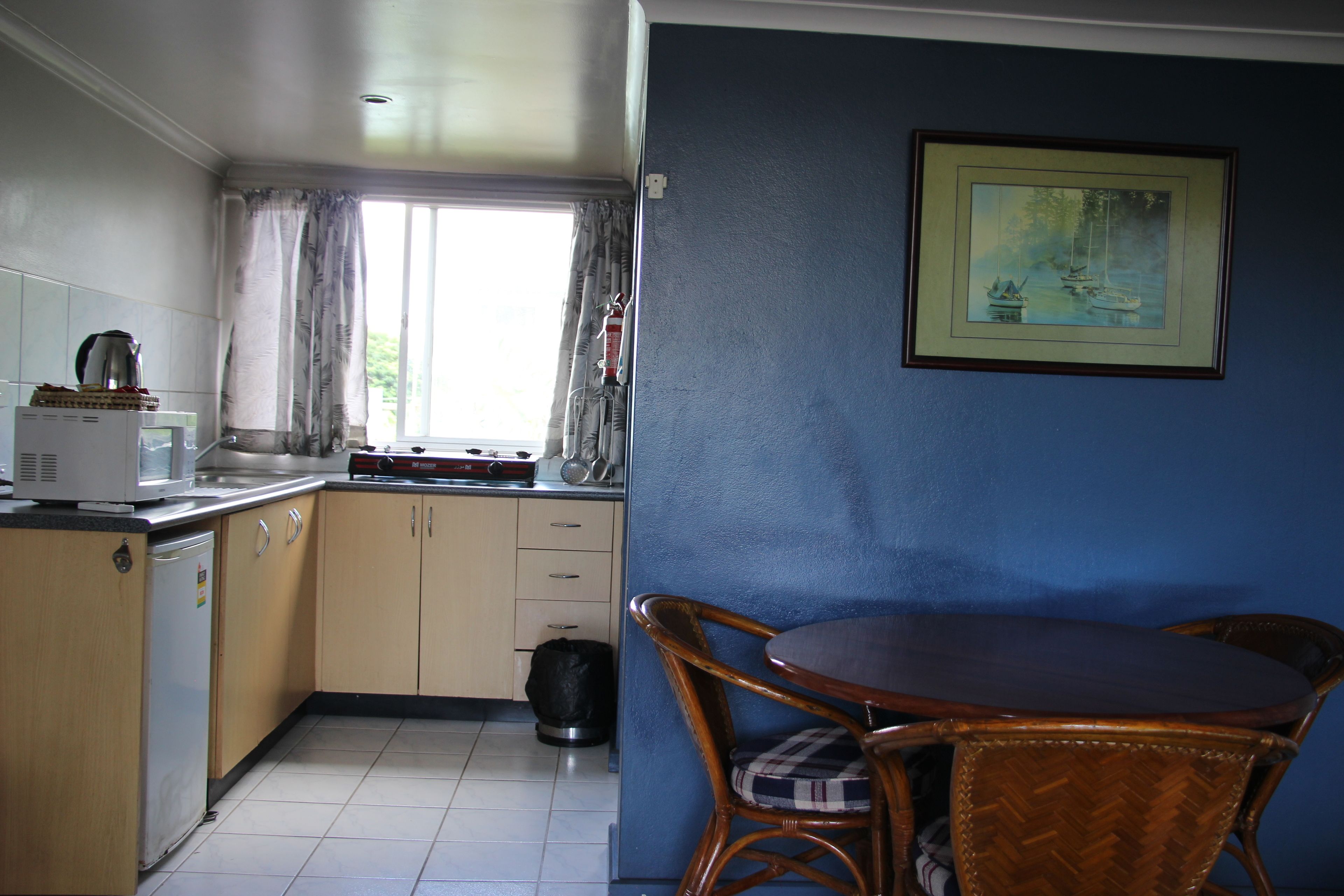 Cozinha compacta com geladeira, micro-ondas e pia. Há uma mesa de jantar com cadeiras e uma janela com cortinas.