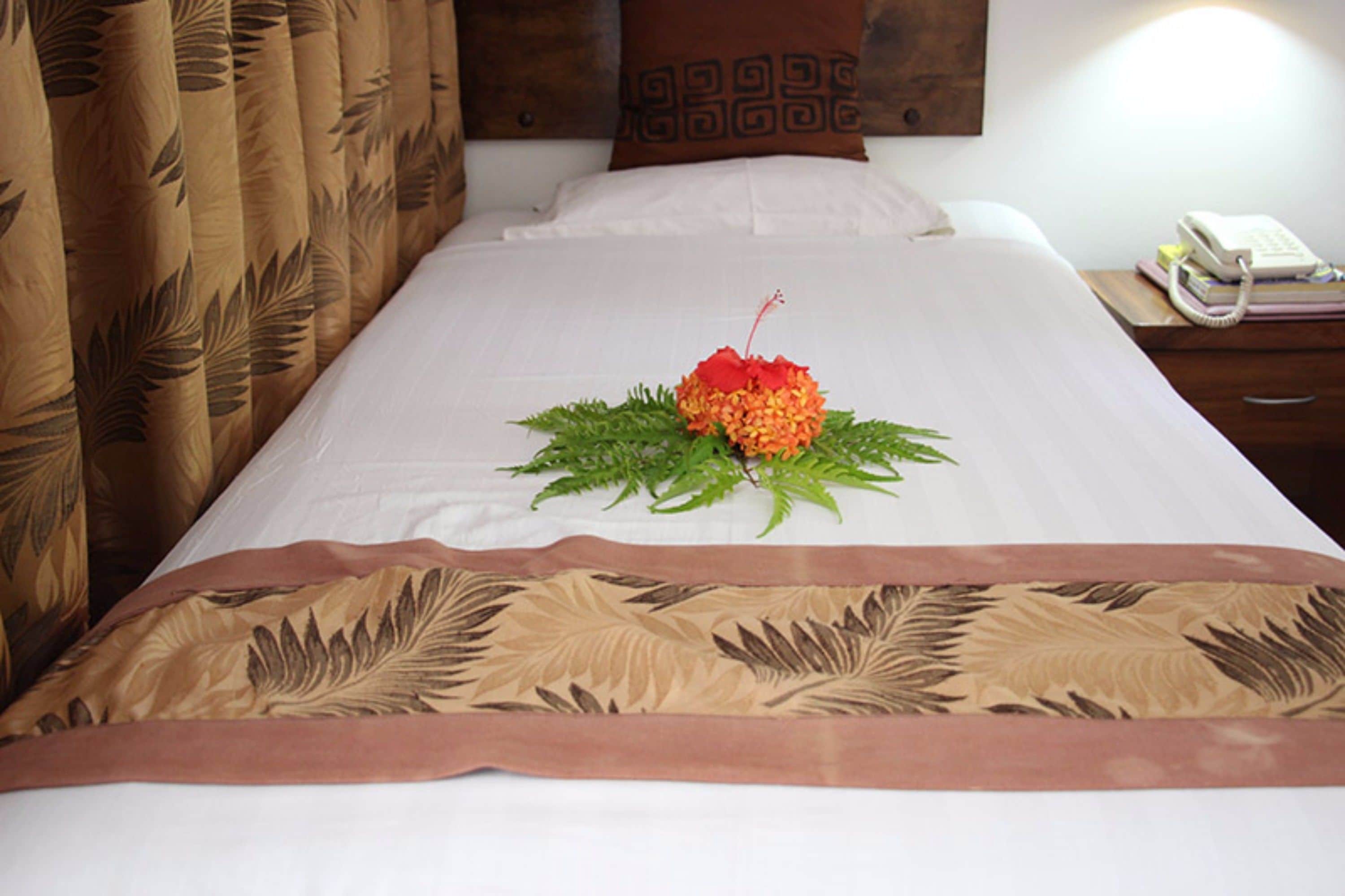 A neatly made bed with a decorative centerpiece, a bedside table with a telephone and books, and a patterned curtain.