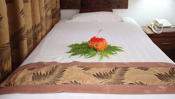 A neatly made bed with a decorative centerpiece, a bedside table with a telephone and books, and a patterned curtain.