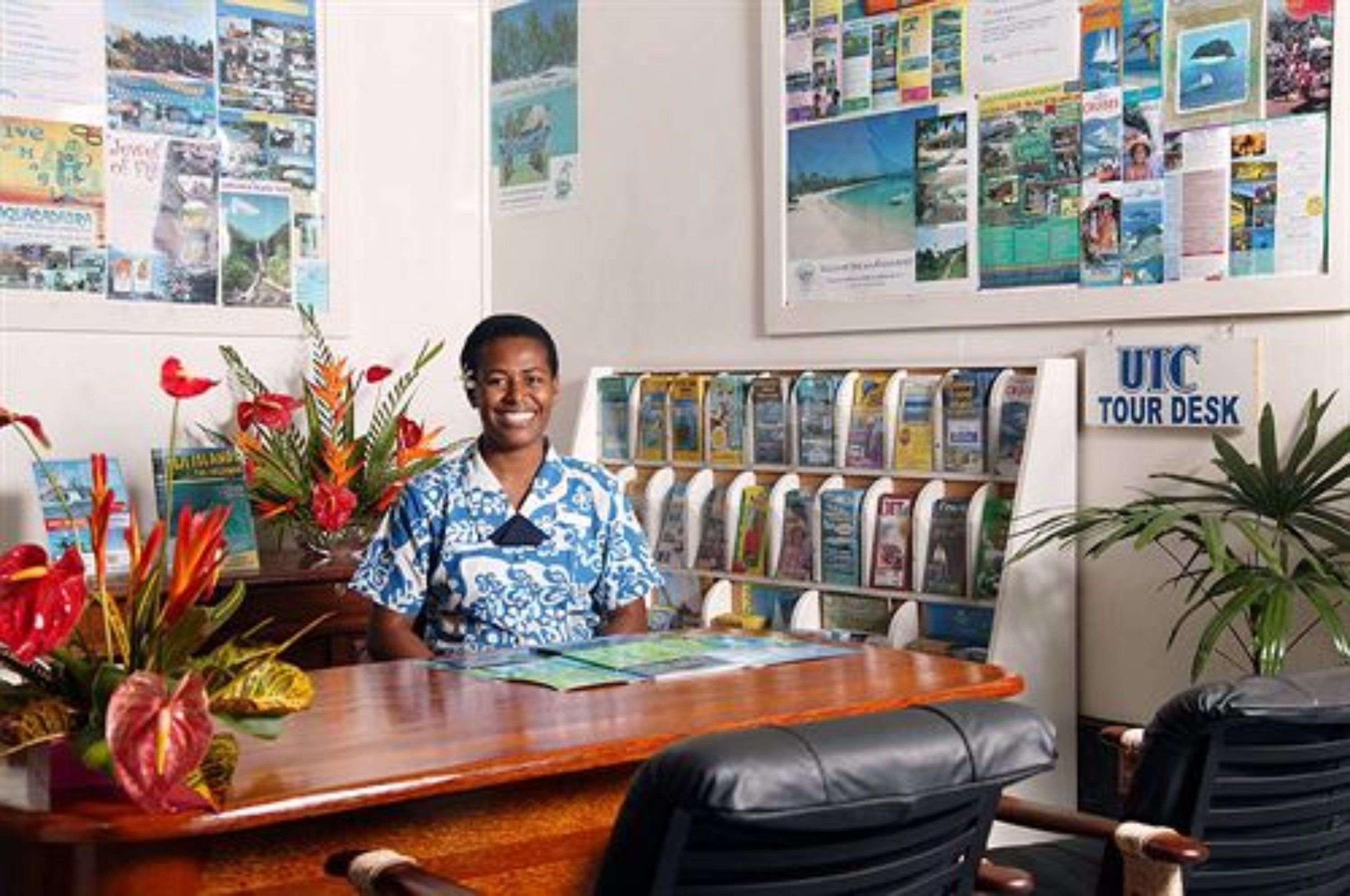 A tour desk with a woman seated behind it, surrounded by travel brochures and a bulletin board with travel photos.