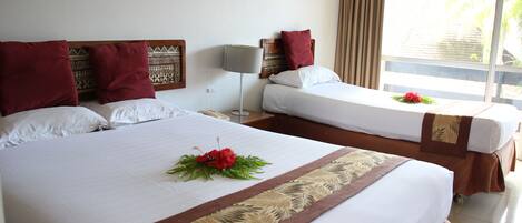 A hotel room with two beds, each with white linens and red pillows. There is a decorative headboard with a patterned design, a bedside table with a lamp, and a window with curtains.