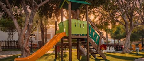 A playground with a slide and climbing structures, surrounded by trees and a paved path.