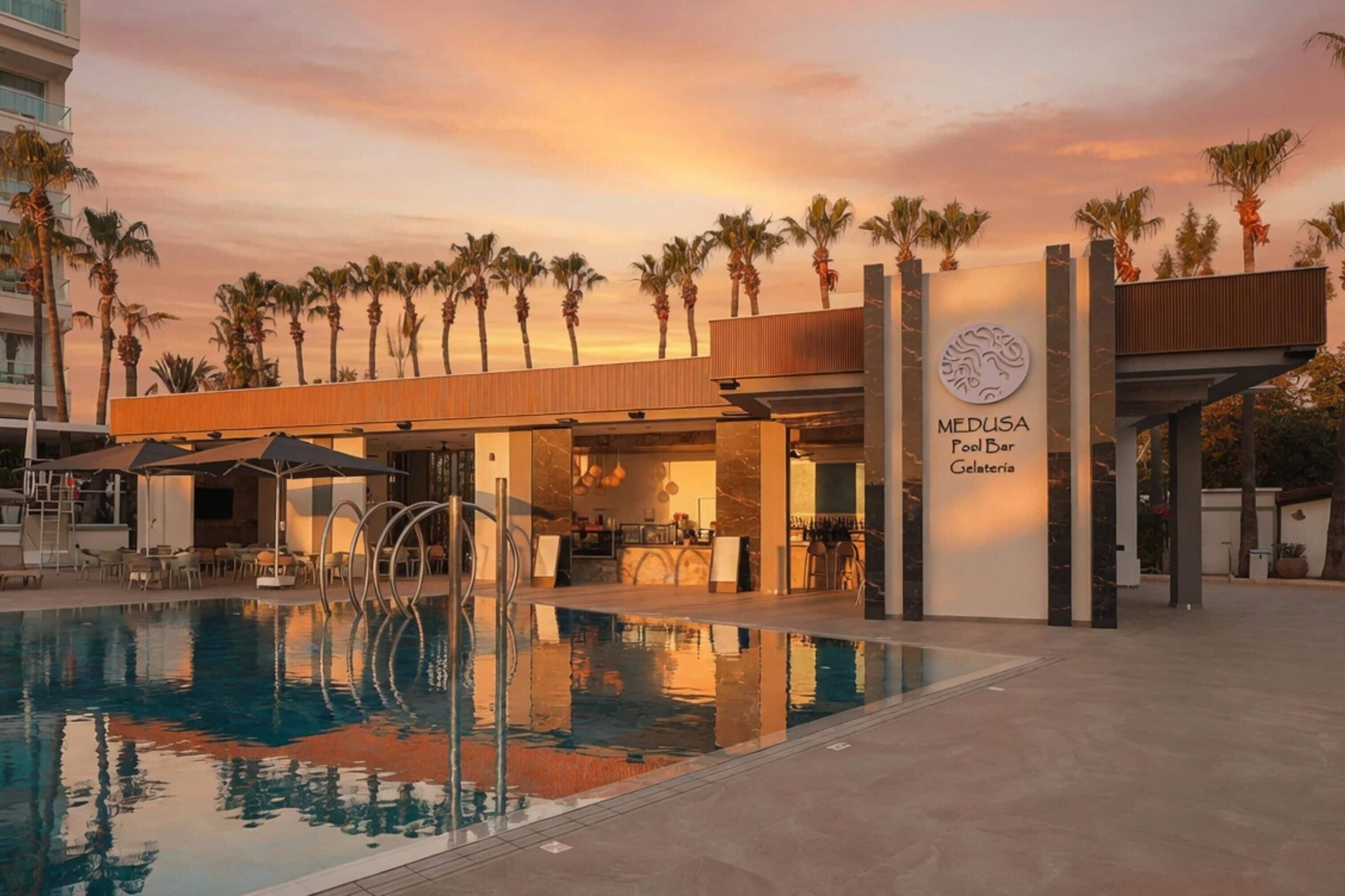 A swimming pool with a bar and restaurant, palm trees, and a sunset sky.
