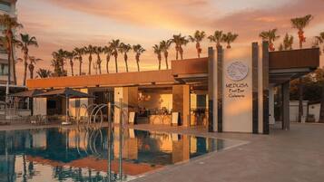 A swimming pool with a bar and restaurant, palm trees, and a sunset sky.