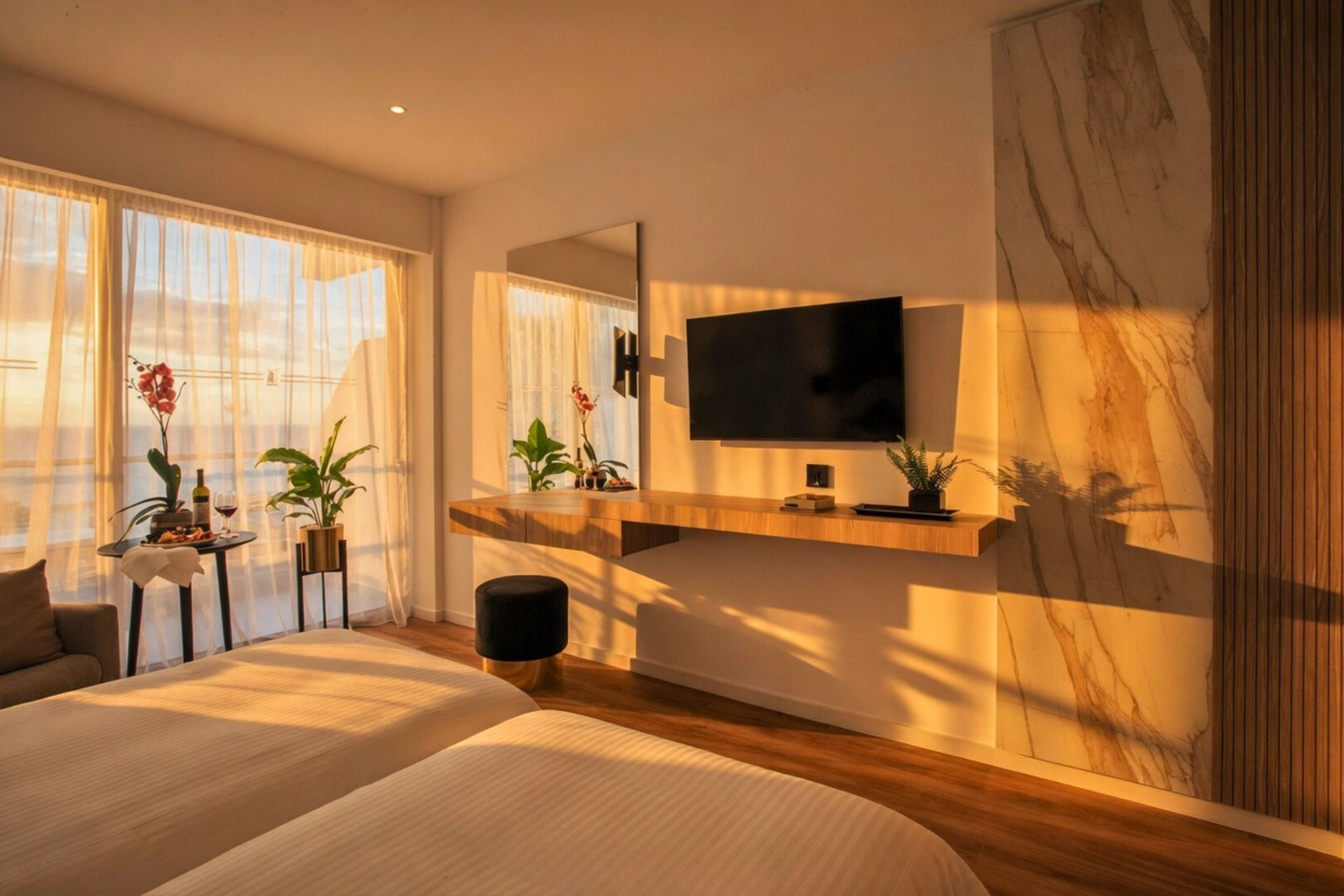 A modern hotel room with a bed, a TV mounted on the wall, a small table with a plant, and a large window with curtains.