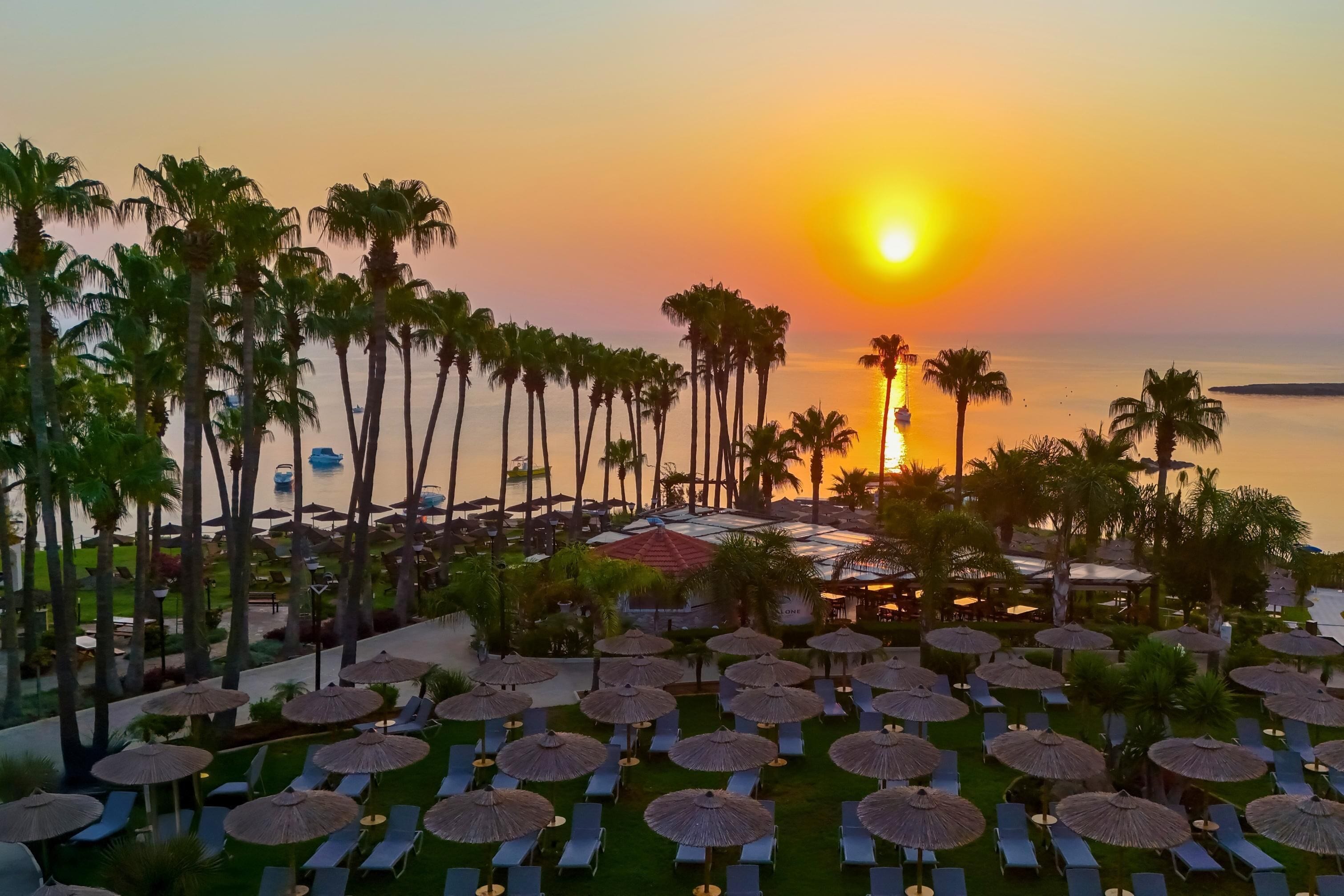 Un complexe hôtelier doté de chaises longues et de parasols, de palmiers et offrant une vue imprenable sur le coucher de soleil.