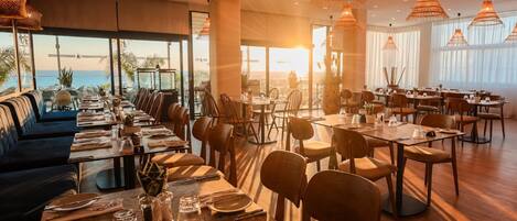 A restaurant with multiple dining tables and chairs, large windows with a view, and hanging pendant lights.