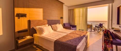 A hotel room with a large bed, a desk, a chair, a wheelchair, and a view of the sea.